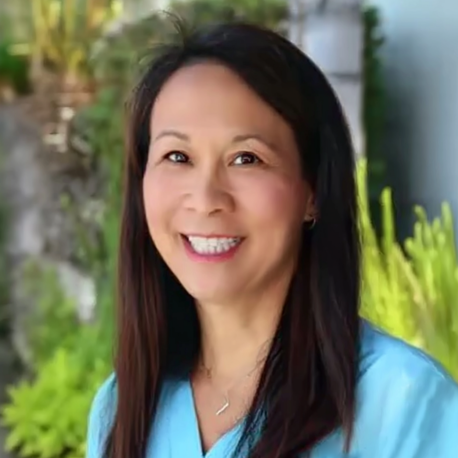 Smiling woman with long dark hair in a light blue top, standing outdoors with greenery behind her