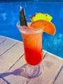 Red tropical cocktail with orange garnish and pineapple on a poolside table.