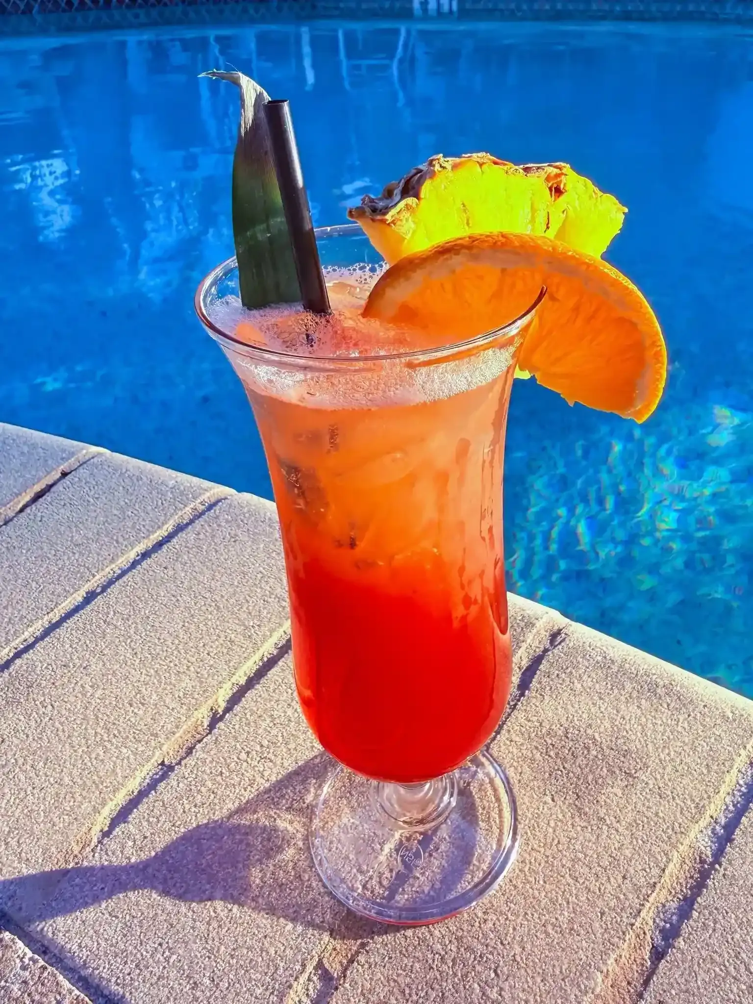 Red tropical cocktail with orange garnish and pineapple on a poolside table.