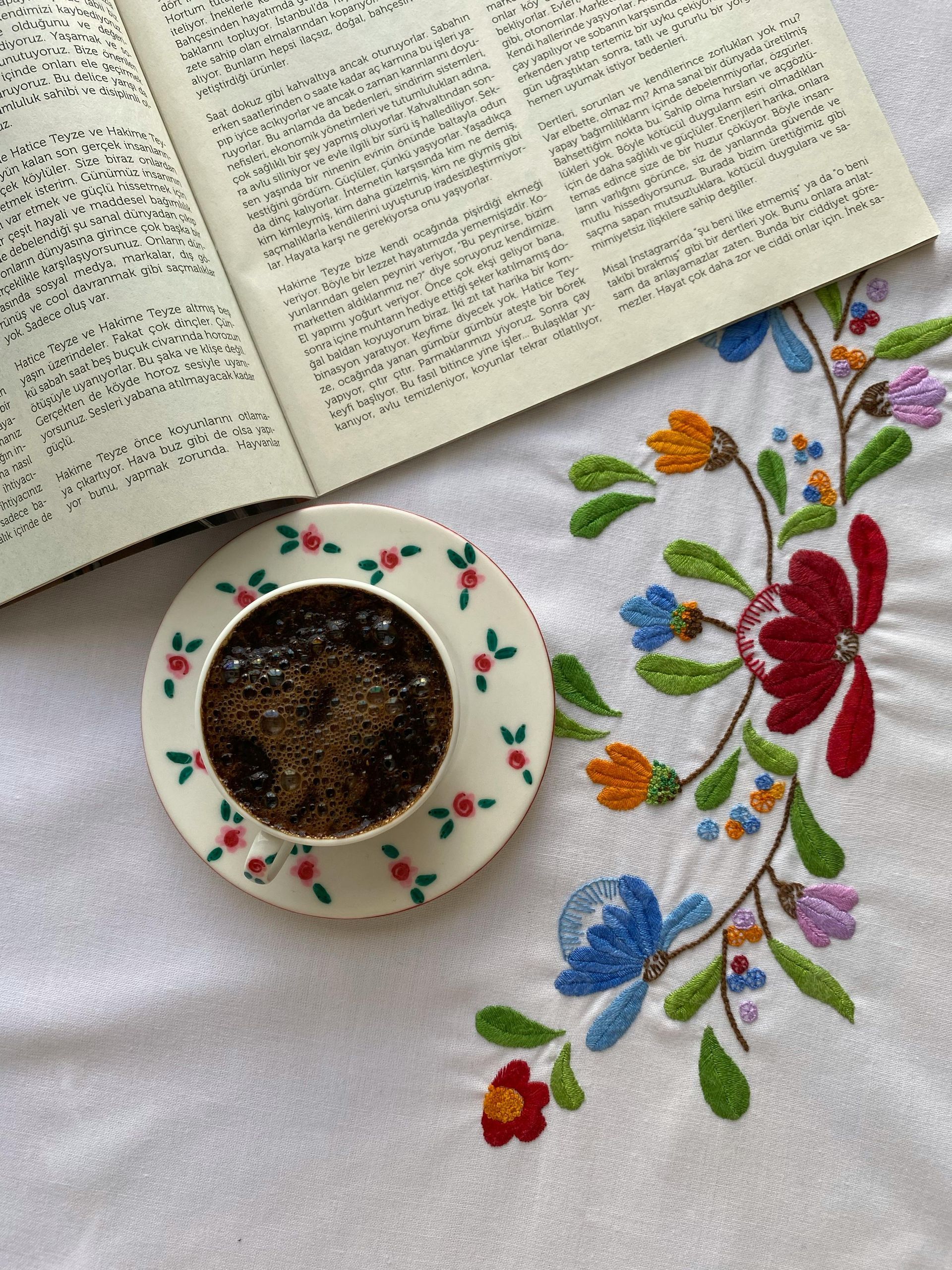 A cup of dark coffee sits on a floral saucer next to an open book, resting on a white tablecloth with embroidered flowers.