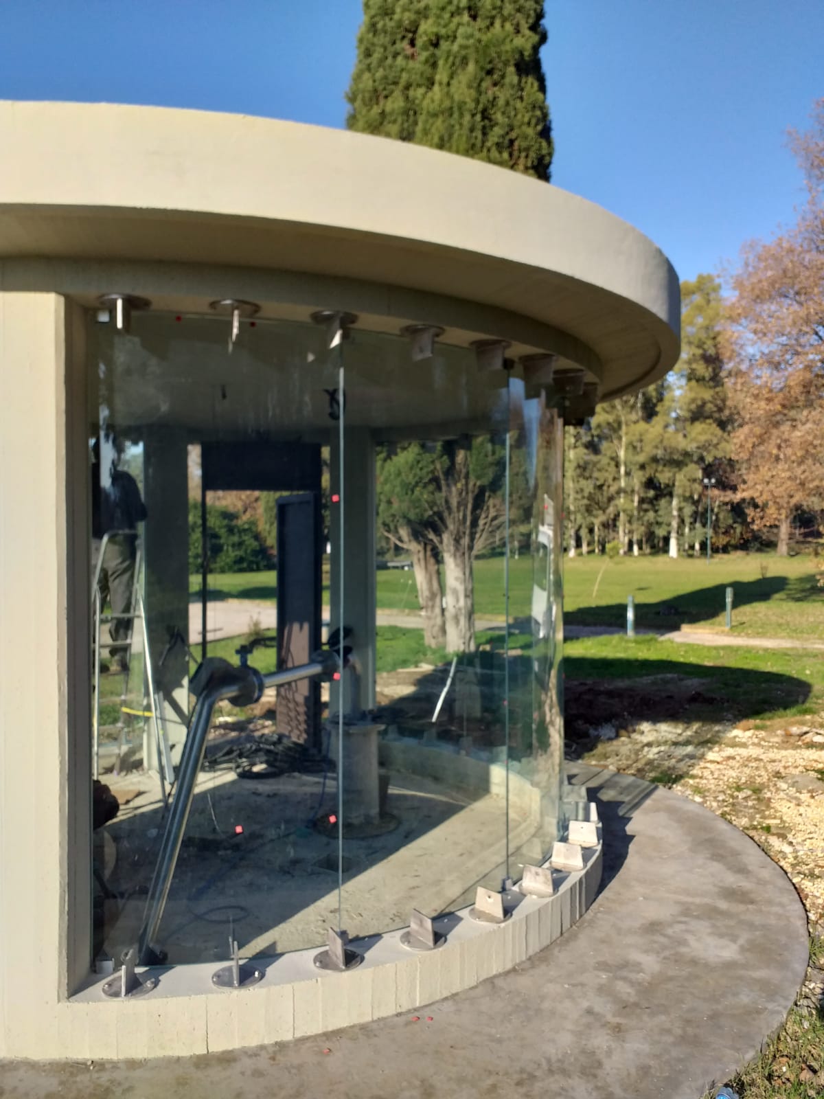 Padiglione moderno con pareti in vetro, attrezzature per l'esercizio fisico, tetto beige curvo e alberi in un parco.