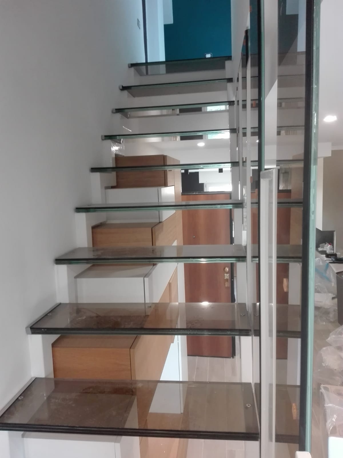 Scala moderna in vetro con gradini in legno accanto a una parete a specchio in un interno luminoso.