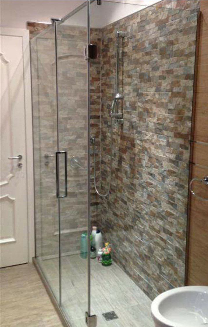 Doccia moderna in bagno con porta in vetro, parete rivestita con piastrelle a mosaico e lavabo bianco in primo piano.