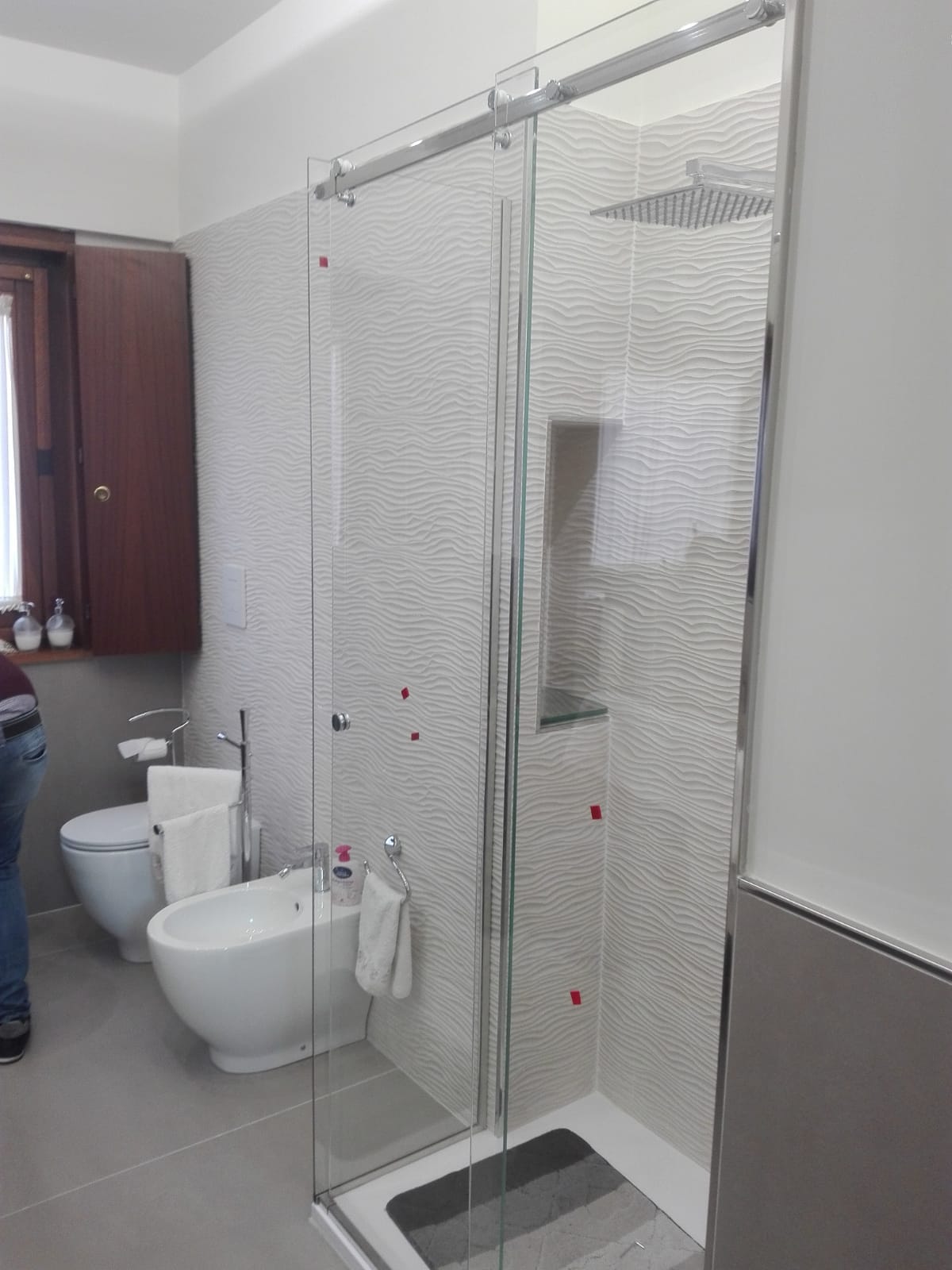 Bagno pubblico con cabine WC bianche, orinatoio e zona doccia separata da pareti in vetro.