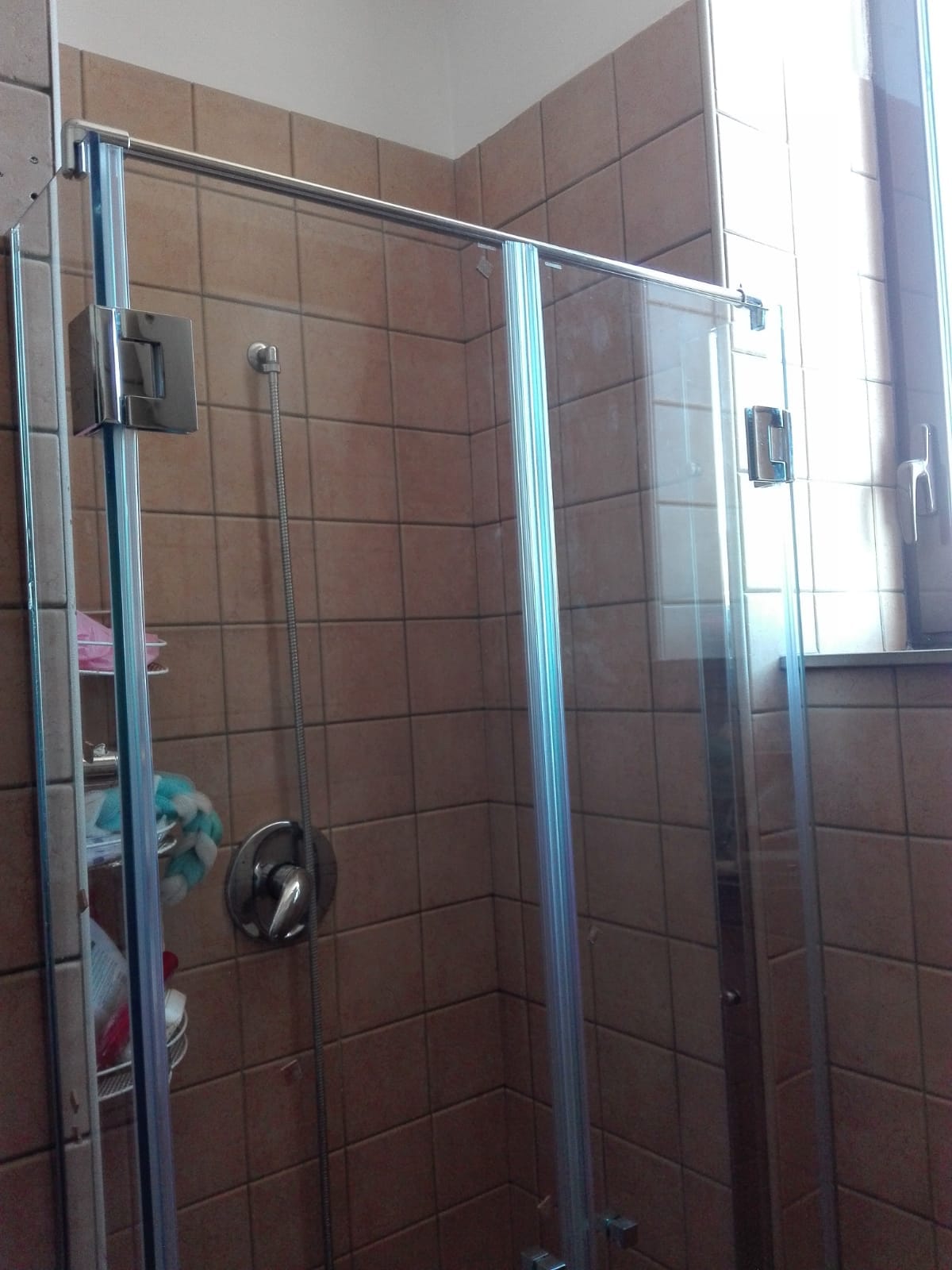 Box doccia in vetro in un bagno piastrellato con maniglia cromata e finestra nelle vicinanze.