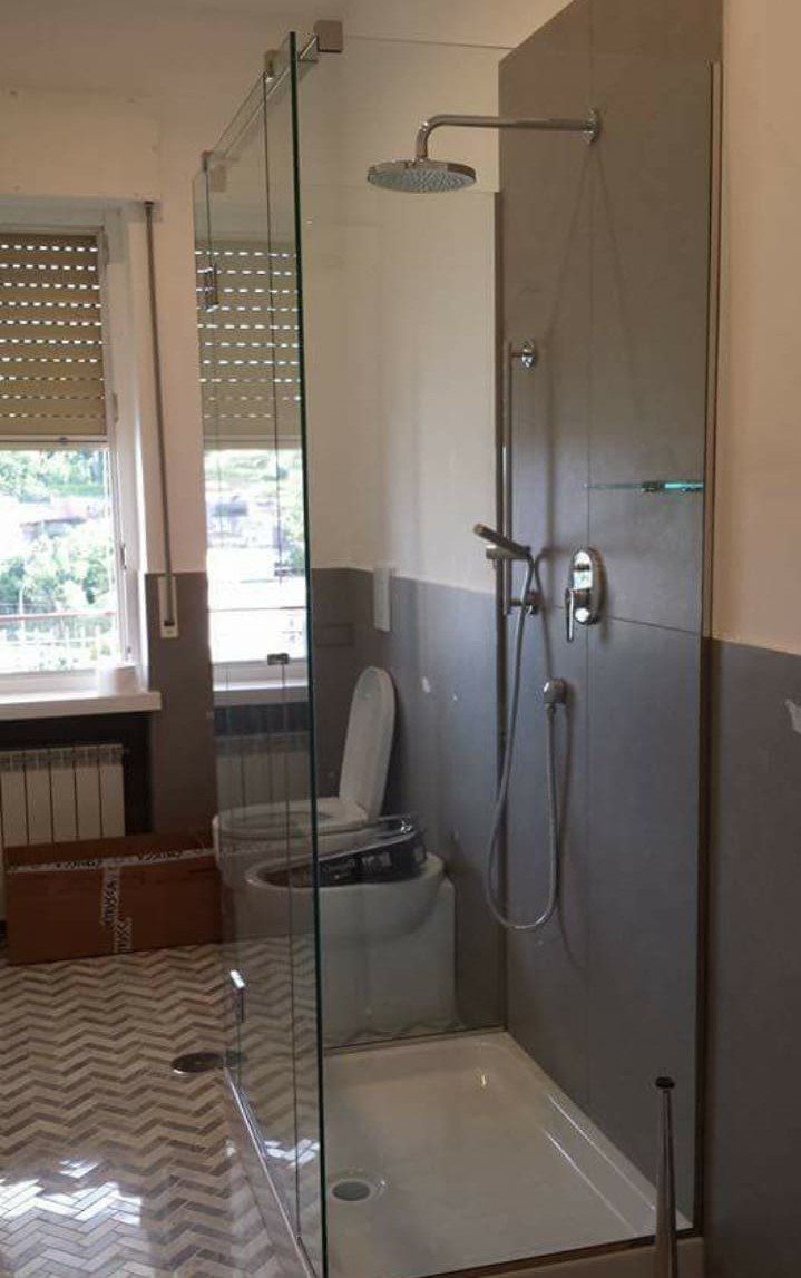 Bagno con box doccia in vetro, doccia a parete, WC e finestra con tende.