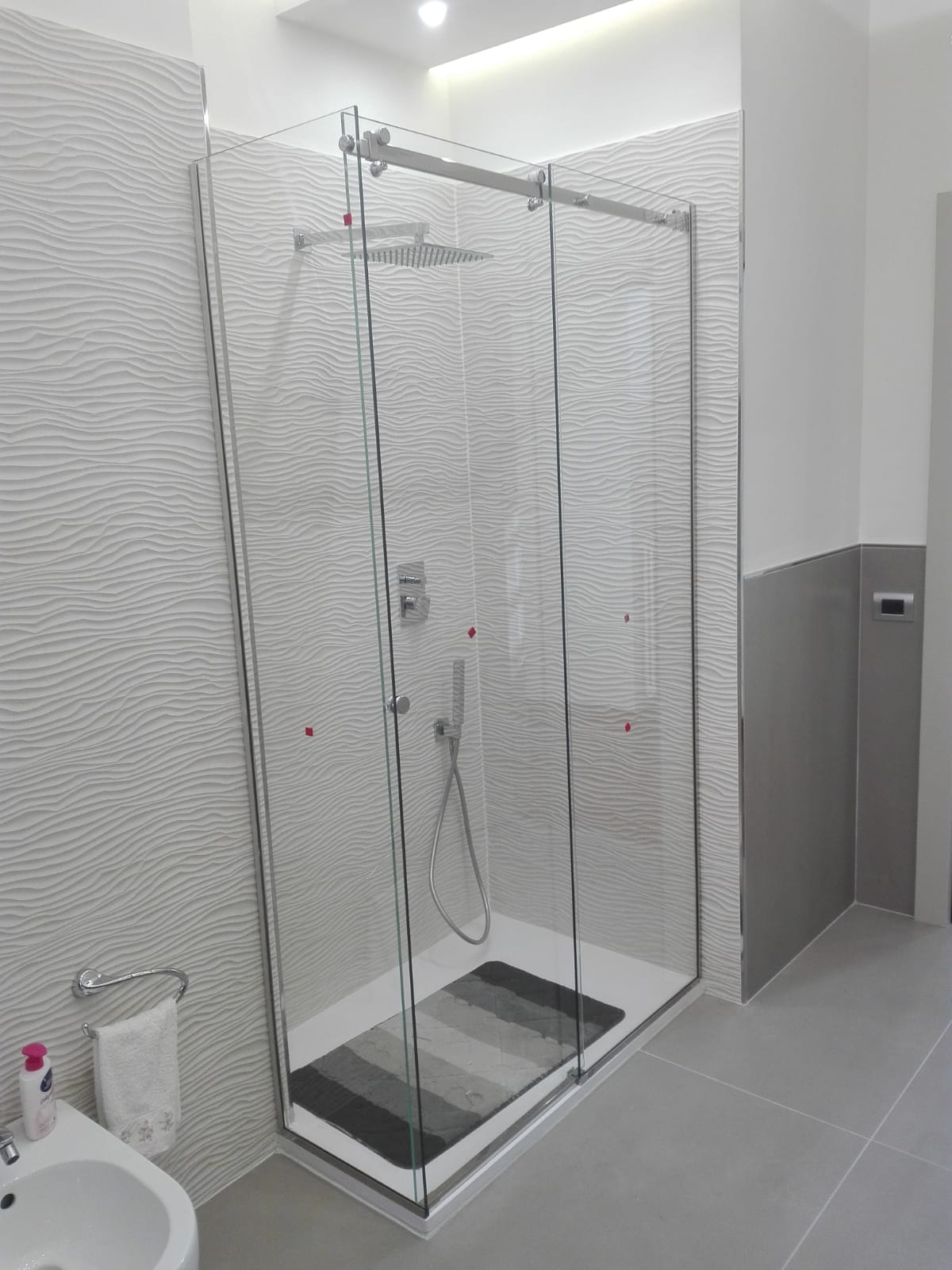 Doccia moderna piastrellata con ante in vetro trasparente, rubinetteria cromata e piatto doccia scuro in un bagno luminoso.