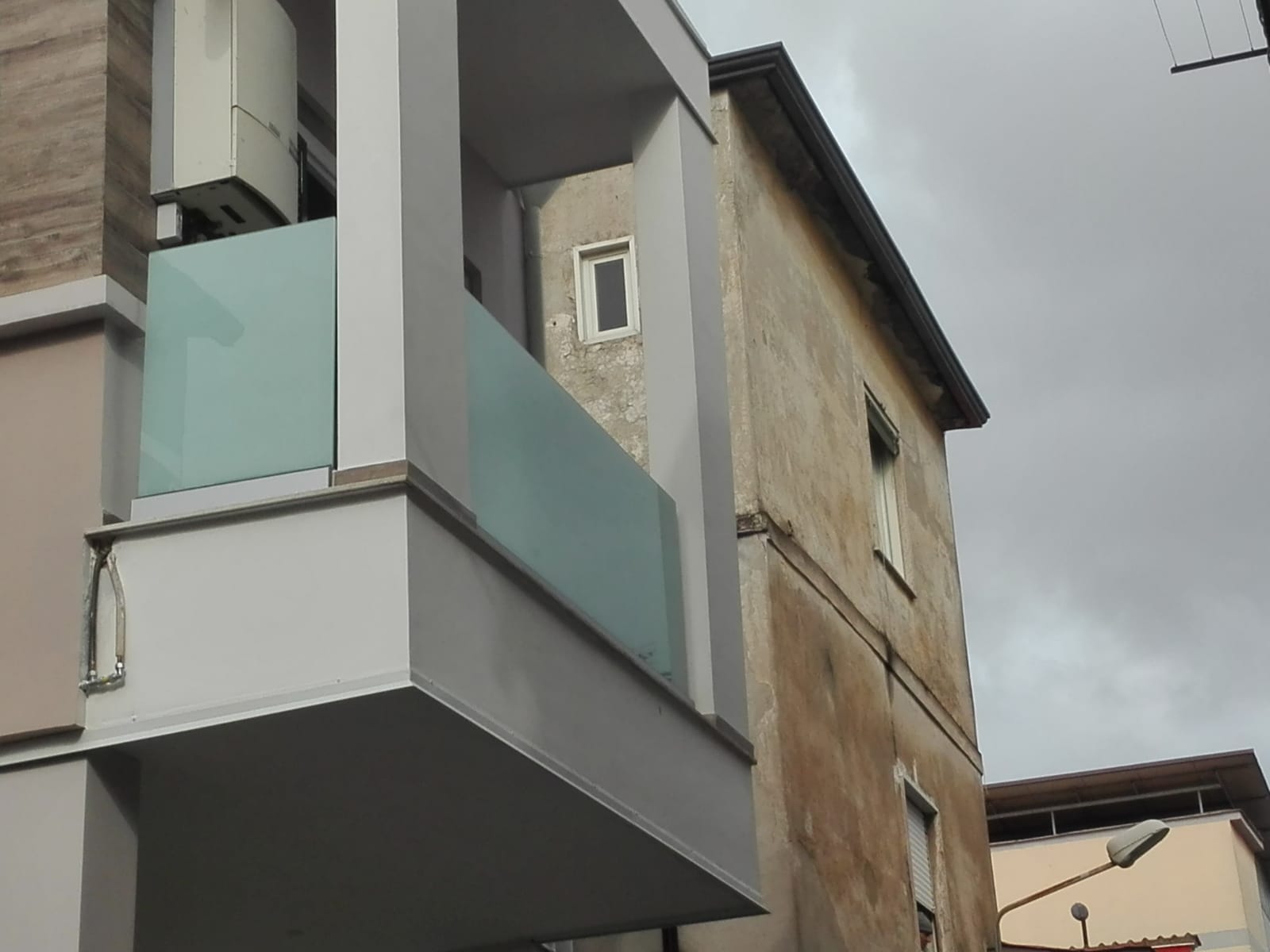Balconi di appartamenti moderni con ringhiere in vetro accanto a un edificio beige sotto un cielo nuvoloso.