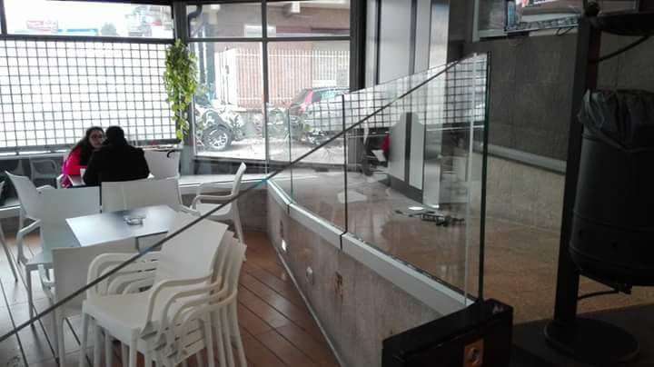 Interno di un caffè moderno con sedie e tavoli bianchi e una parete divisoria in vetro; due persone sono sedute vicino alla finestra.