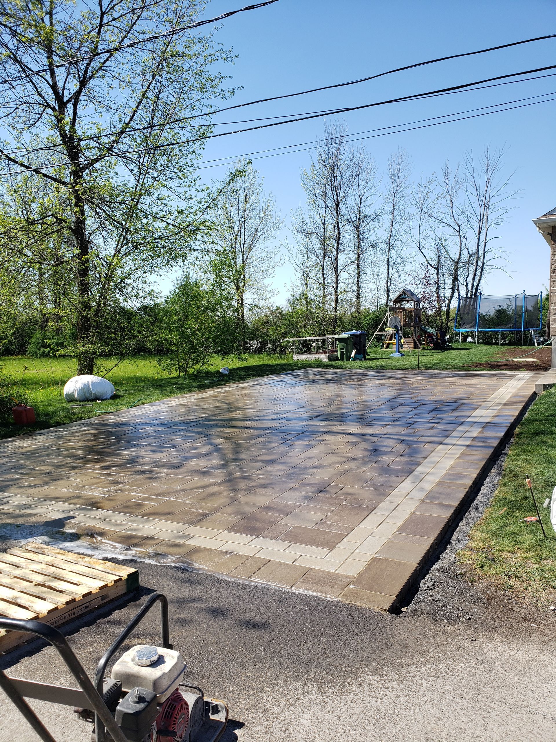 Terrasse en briques fraîchement coulées, arrosée ; un compacteur est stationné à son bord. Arbres verts et ciel d'un bleu limpide.