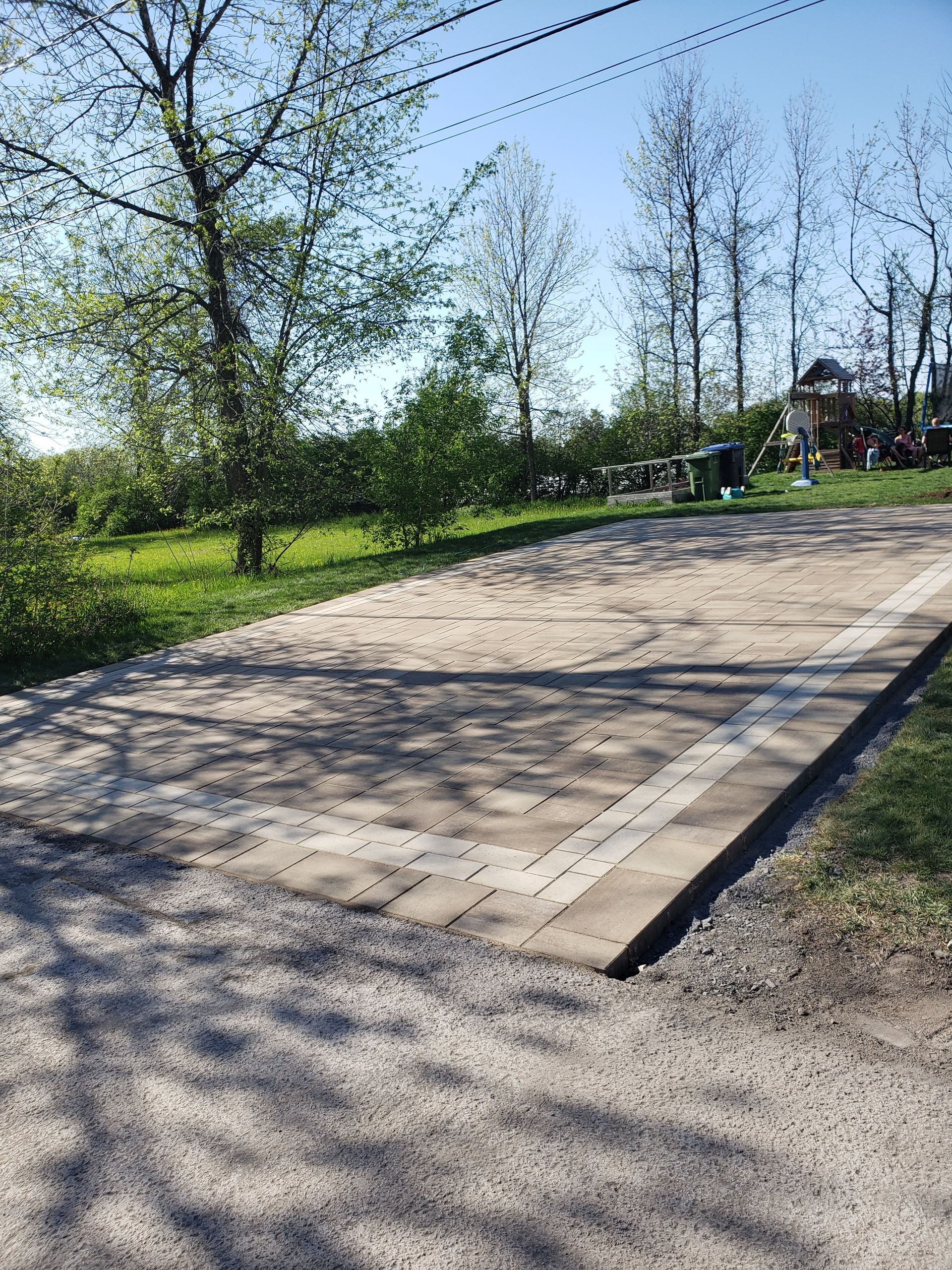 Terrain de pétanque extérieur entouré de gravier, bordé de pavés clairs, d'une pelouse et d'arbres.