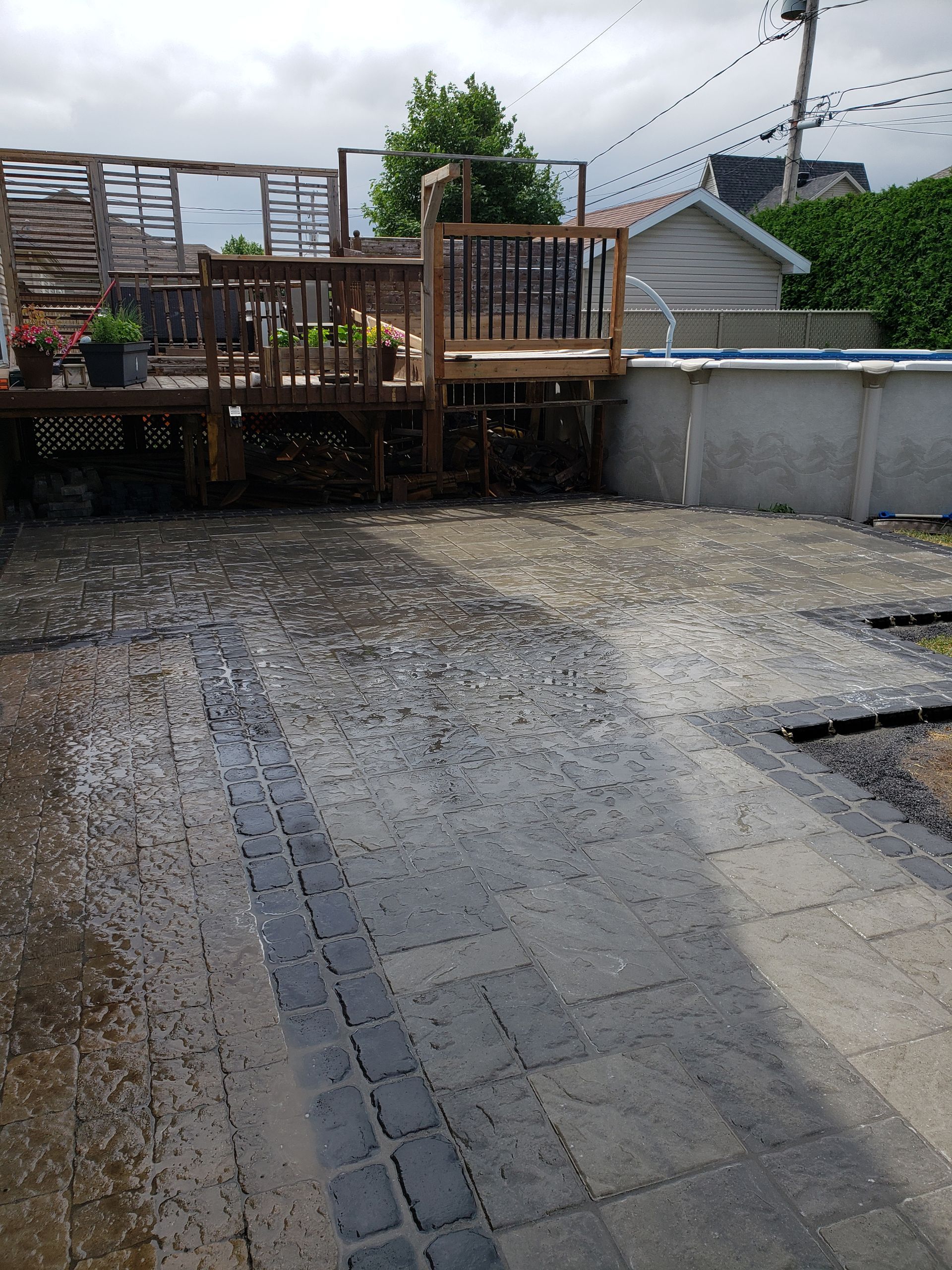 Pluie tombant sur une terrasse pavée avec une terrasse en bois et une piscine hors sol en arrière-plan.
