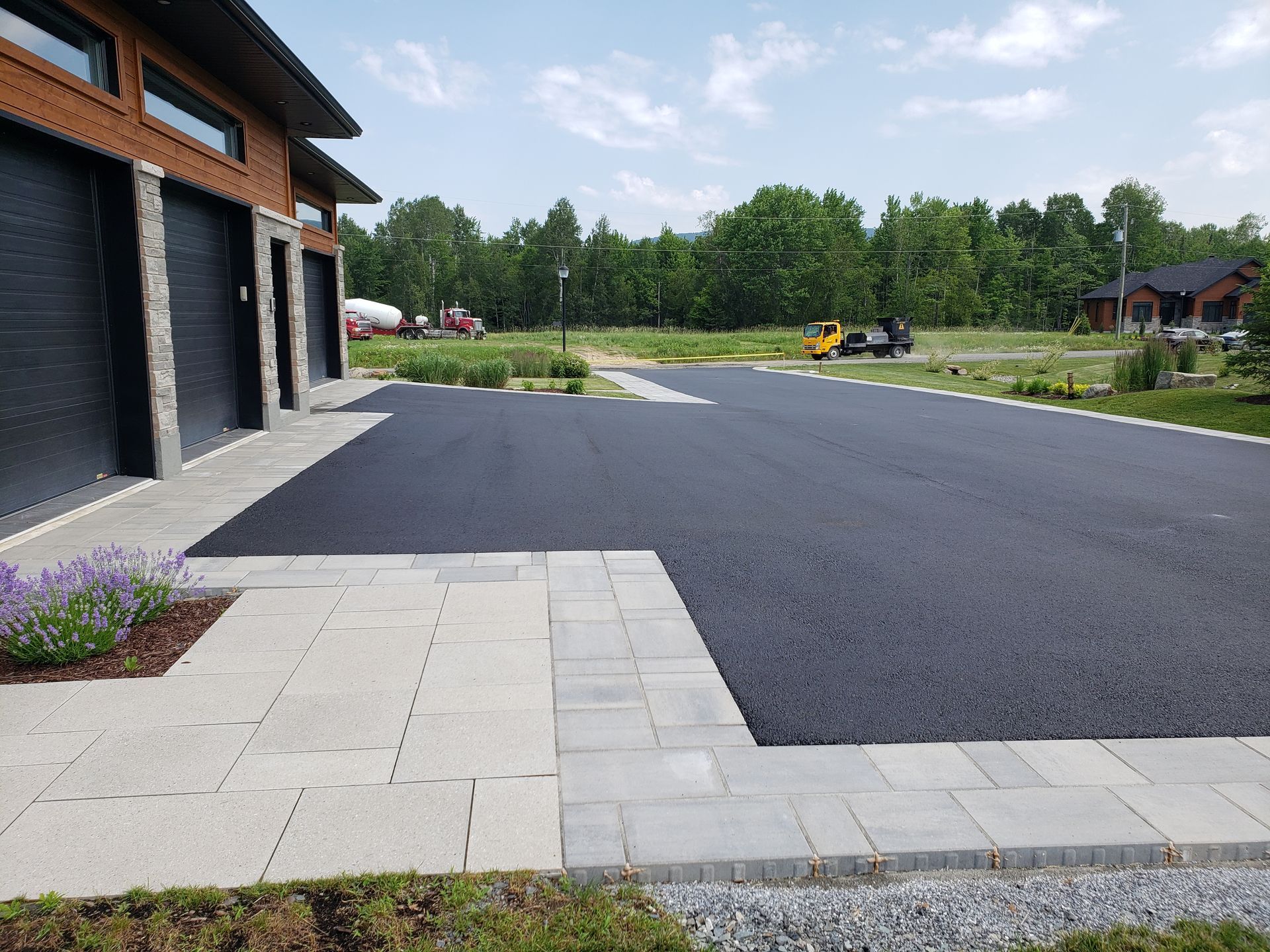 Allée en asphalte noir jouxtant un chemin piétonnier en pierre gris clair et un garage aux portes noires. Aménagement paysager verdoyant à l'avant.