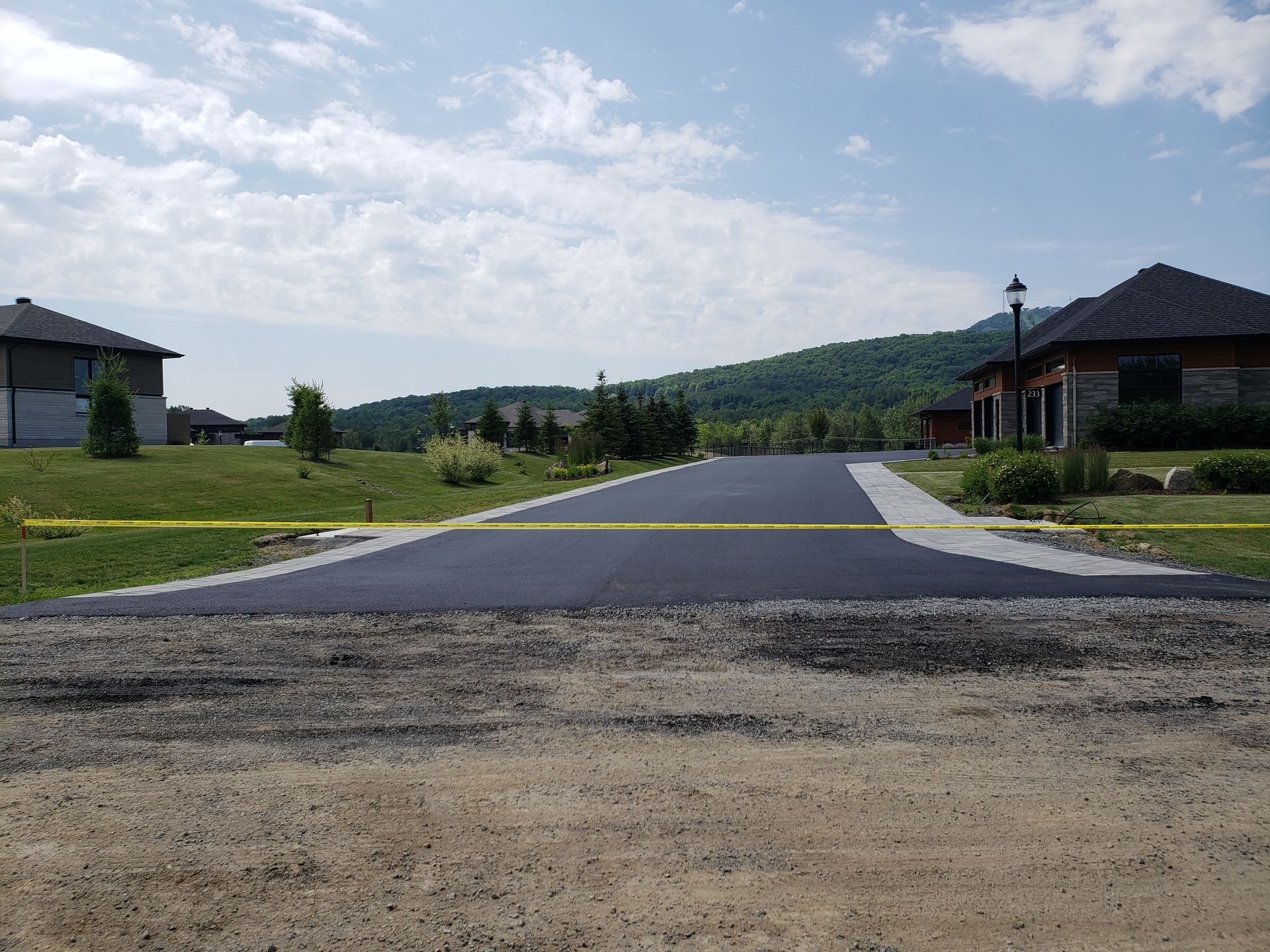Route nouvellement asphaltée bordée de maisons, de ruban de signalisation jaune et d'une montagne en arrière-plan.