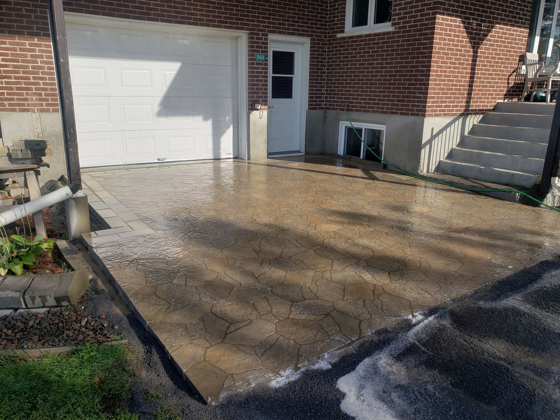 Allée en béton mouillée devant un bâtiment en briques avec un garage, une porte et un escalier menant à l'étage.
