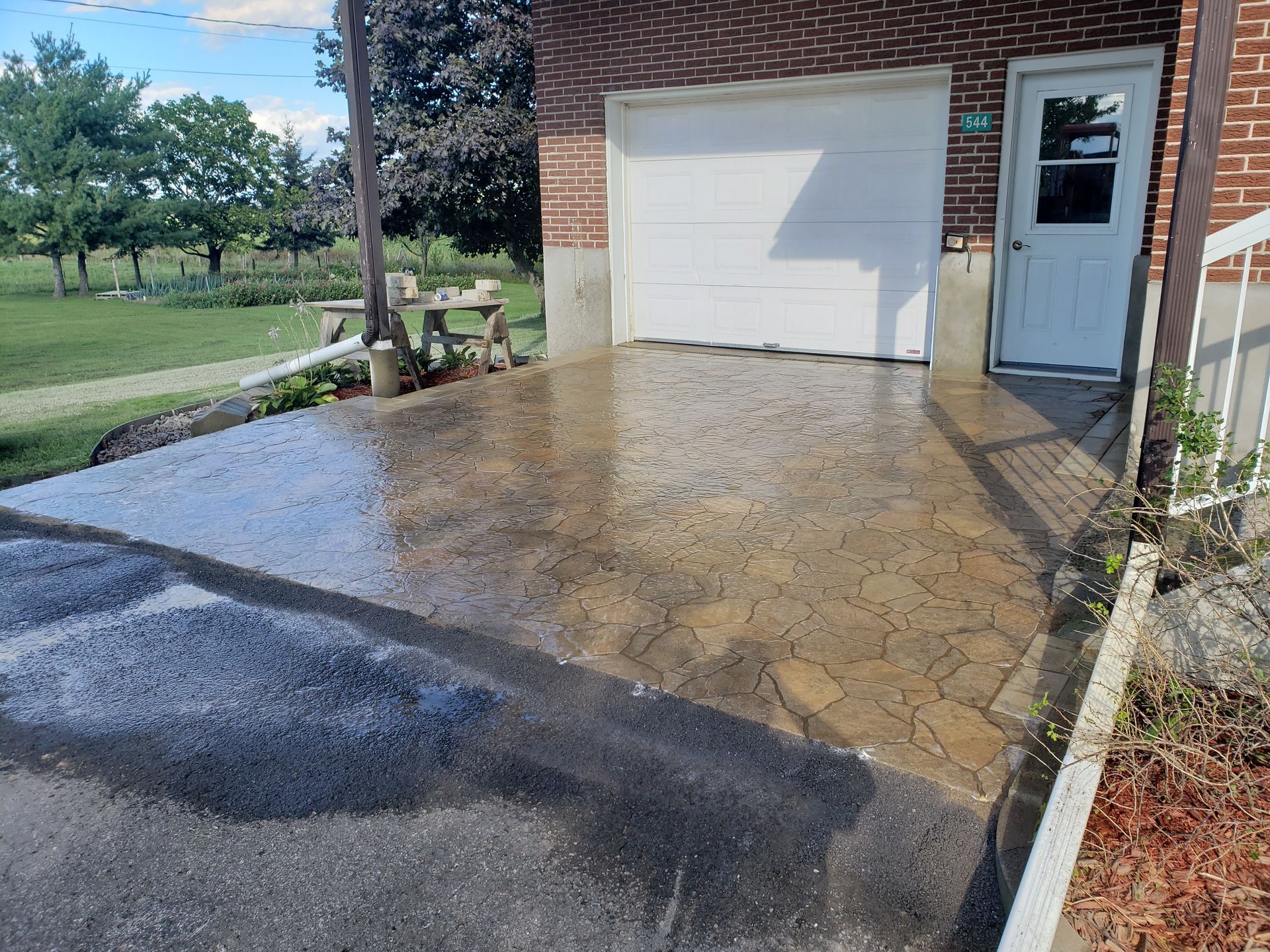 Allée mouillée devant un bâtiment en briques avec une porte de garage et une porte blanche, pelouse sur le côté.