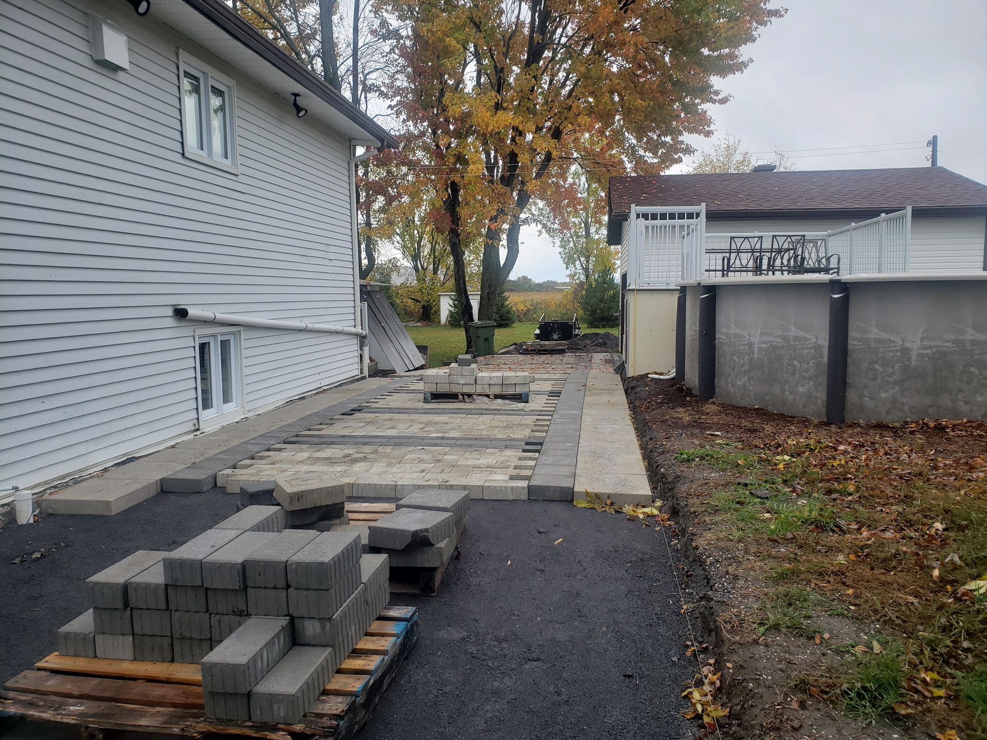 Chantier de construction dans le jardin : maison, dalles de terrasse, piscine, arbres. Briques grises empilées, terrain préparé.