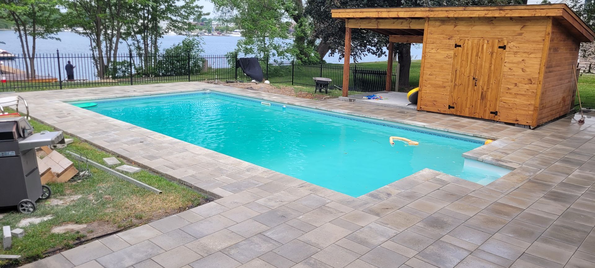 Une piscine dans le jardin, aux eaux turquoise, entourée de dalles de pierre et d'un abri de jardin en bois.