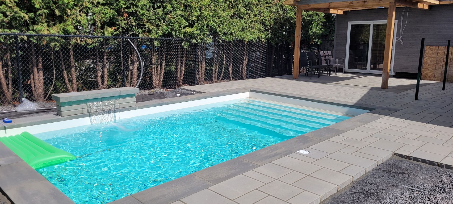 Piscine rectangulaire aux eaux turquoise, entourée de pavés gris et d'une pelouse verte. Clôture noire et abri de jardin en arrière-plan.