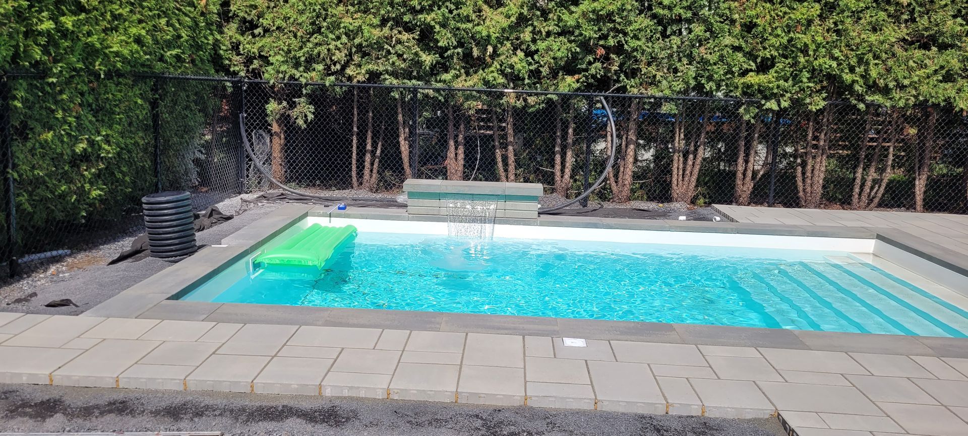 Une piscine rectangulaire turquoise avec un flotteur vert, entourée d'une terrasse pavée et de haies verdoyantes.