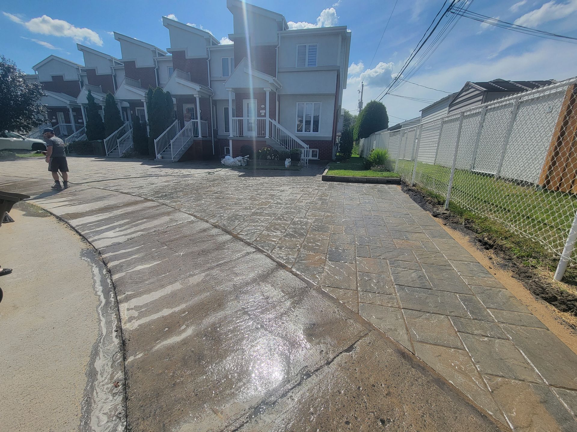 Allée en béton mouillée avec une personne debout sur le côté, maisons en arrière-plan, journée ensoleillée.