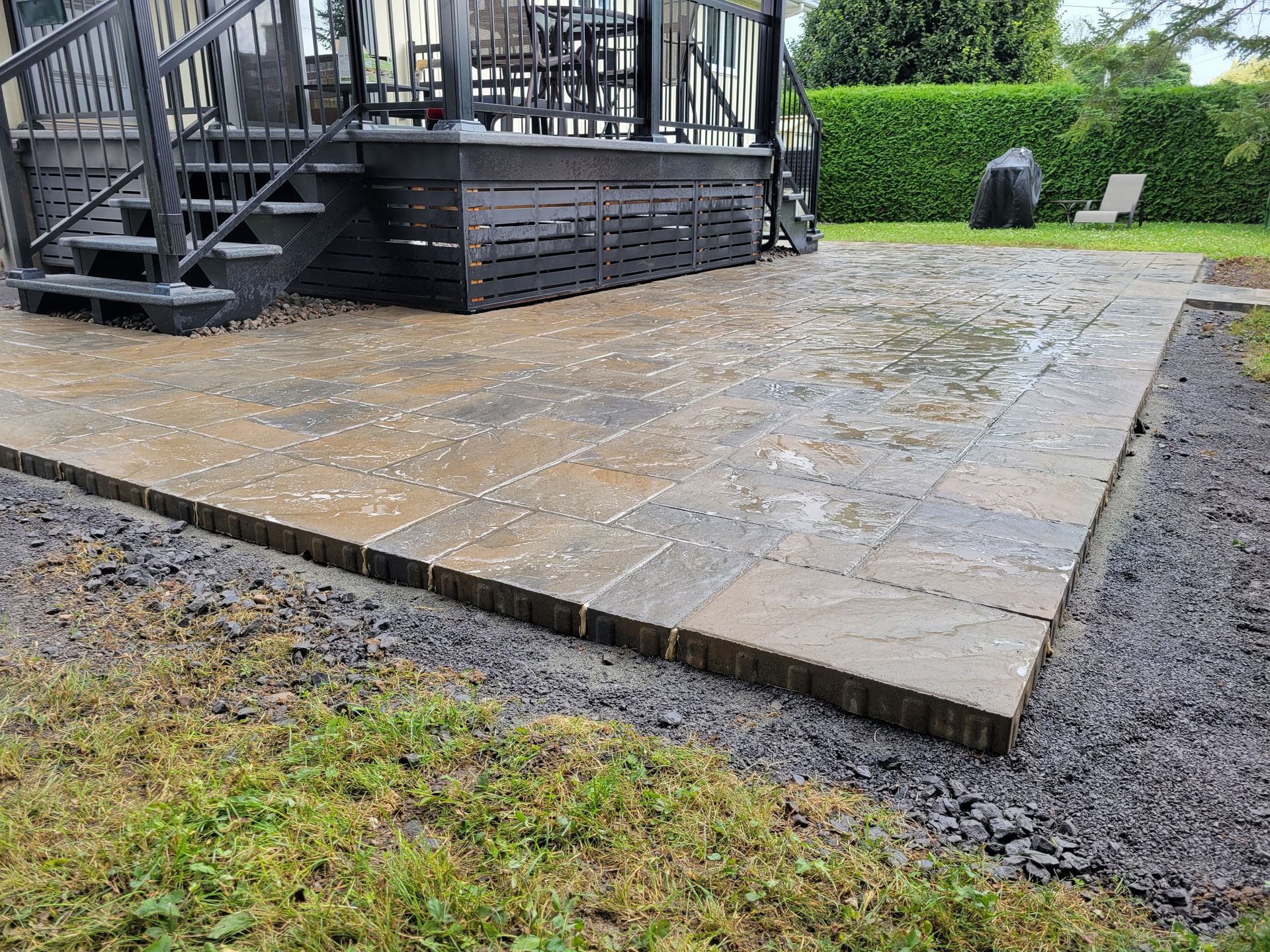 Terrasse pavée de dalles foncées, bordée de blocs foncés, jouxtant une terrasse en bois et une pelouse. Surface humide.