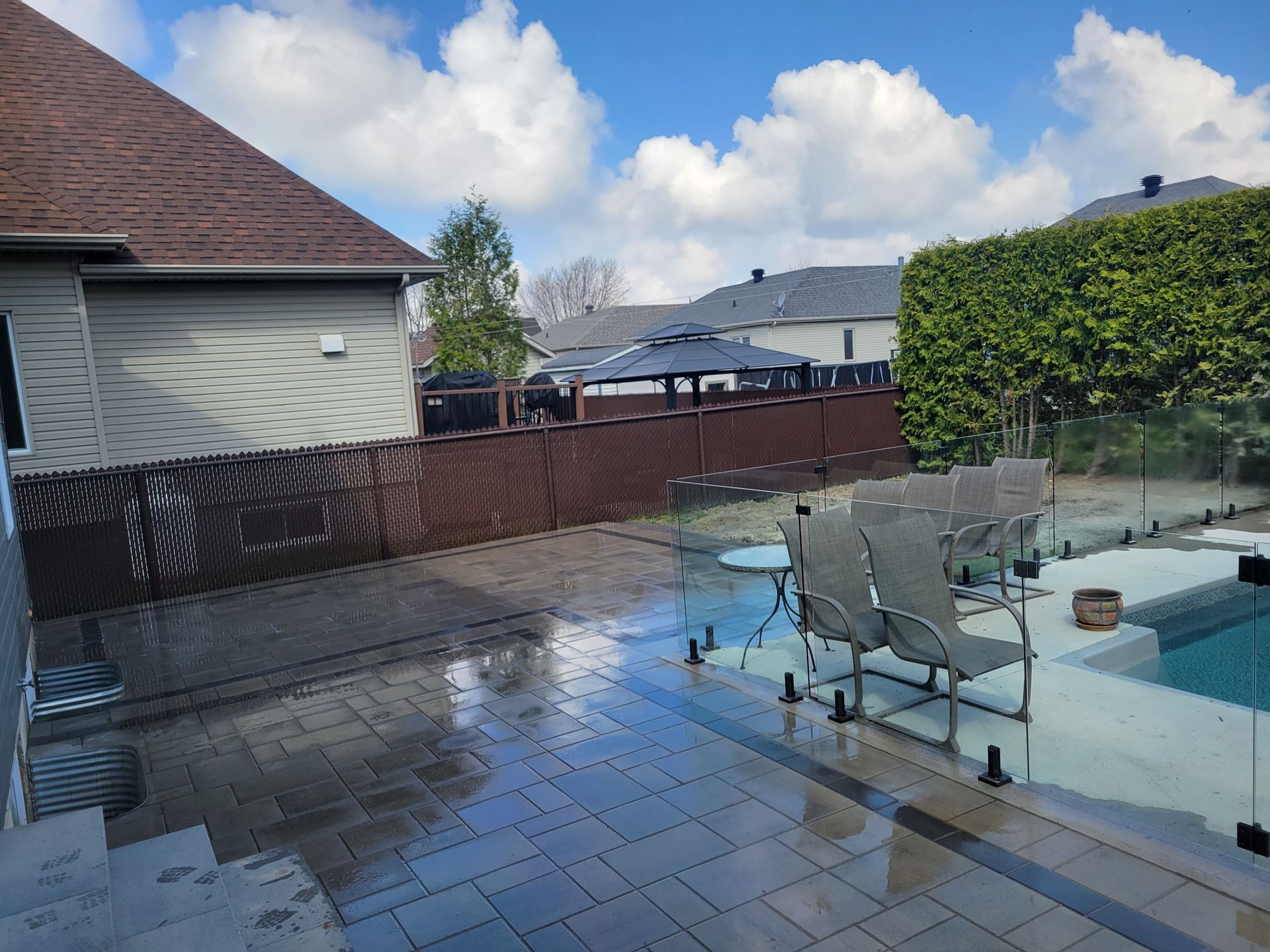 Jardin avec terrasse en pierre humide, piscine, clôture marron et ciel dégagé avec quelques nuages.