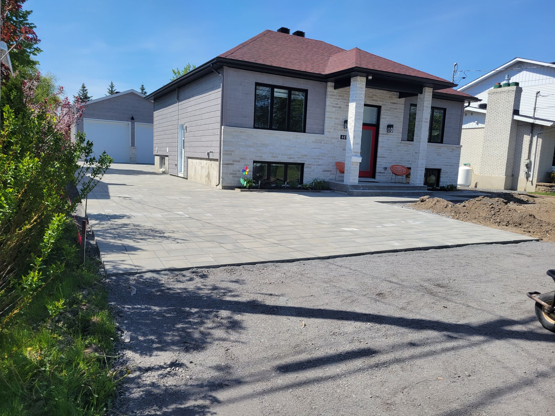 Maison avec allée pavée ; façade grise et blanche, porte rouge, garage en arrière-plan par une journée ensoleillée.