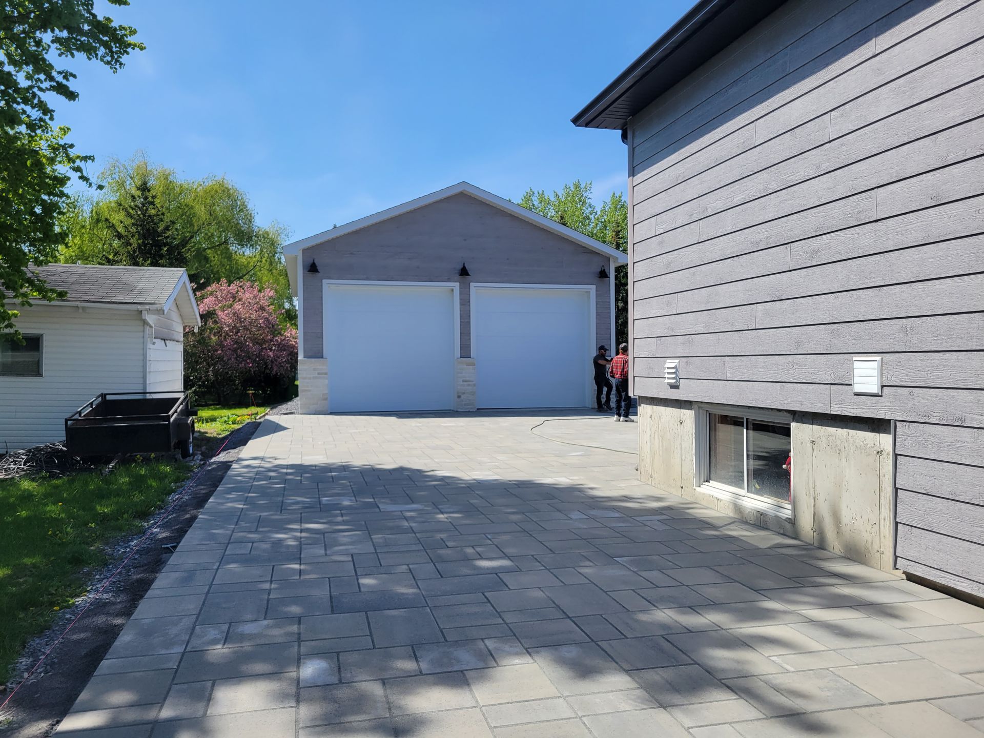 Garage gris pour deux voitures avec portes blanches et soubassement en briques claires, jouxtant une maison grise avec une allée pavée.
