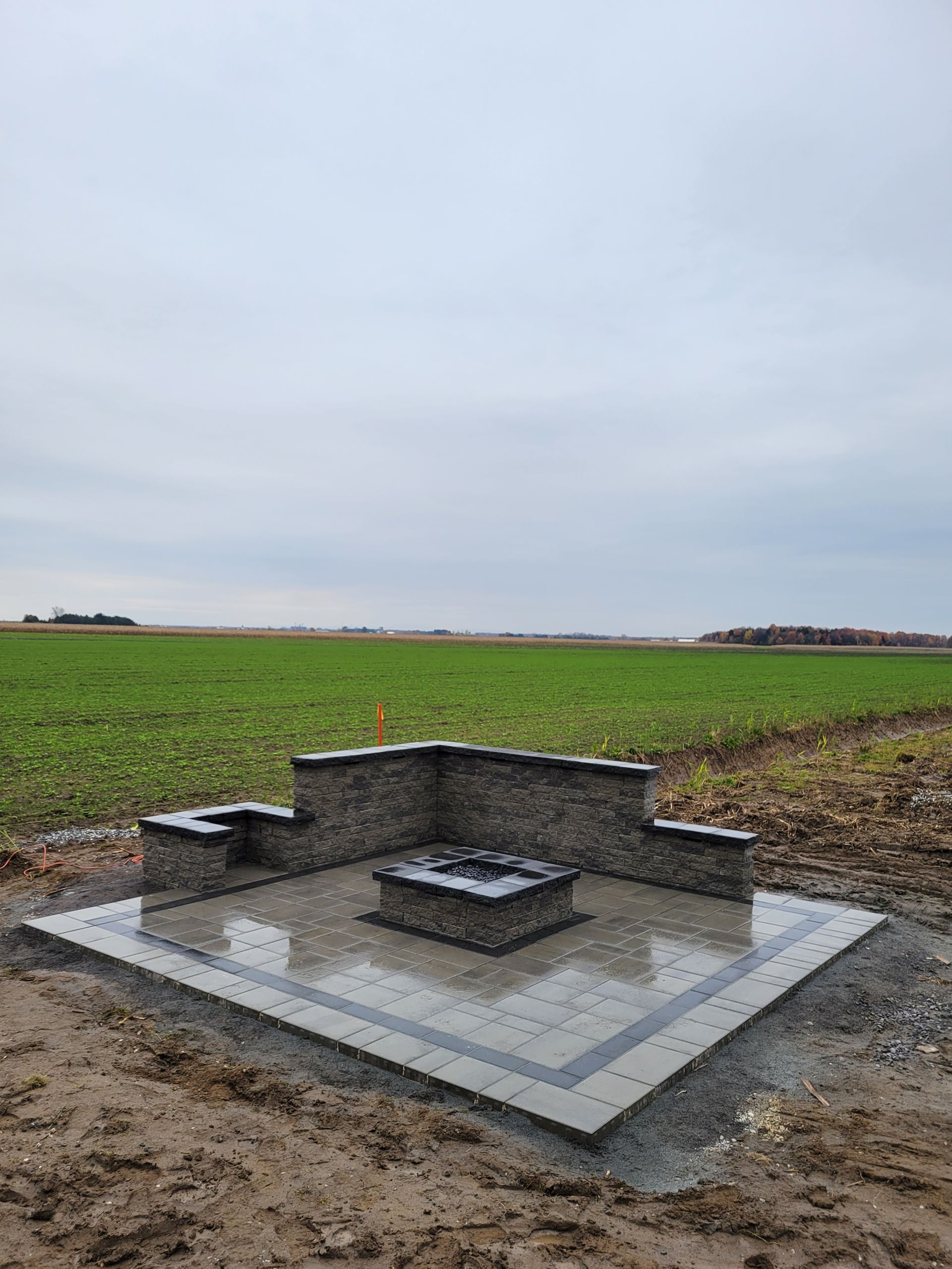 Un foyer en pierre et un coin salon aménagés sur une dalle de béton, dans un champ sous un ciel couvert.
