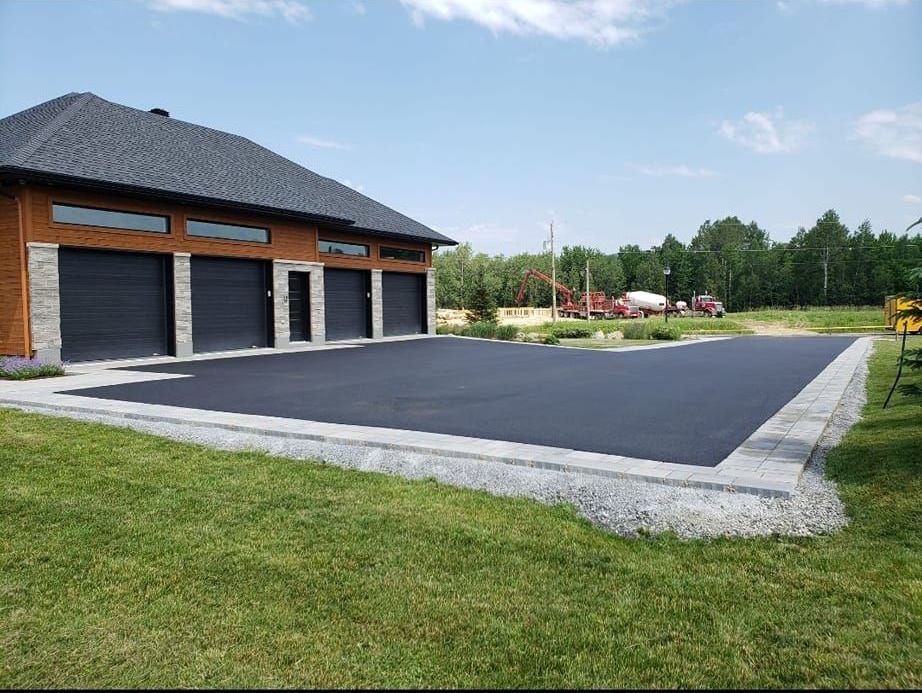 Allée en asphalte noir devant un bâtiment avec portes de garage noires. Bordures en pierre grise. Pelouse verte tout autour.
