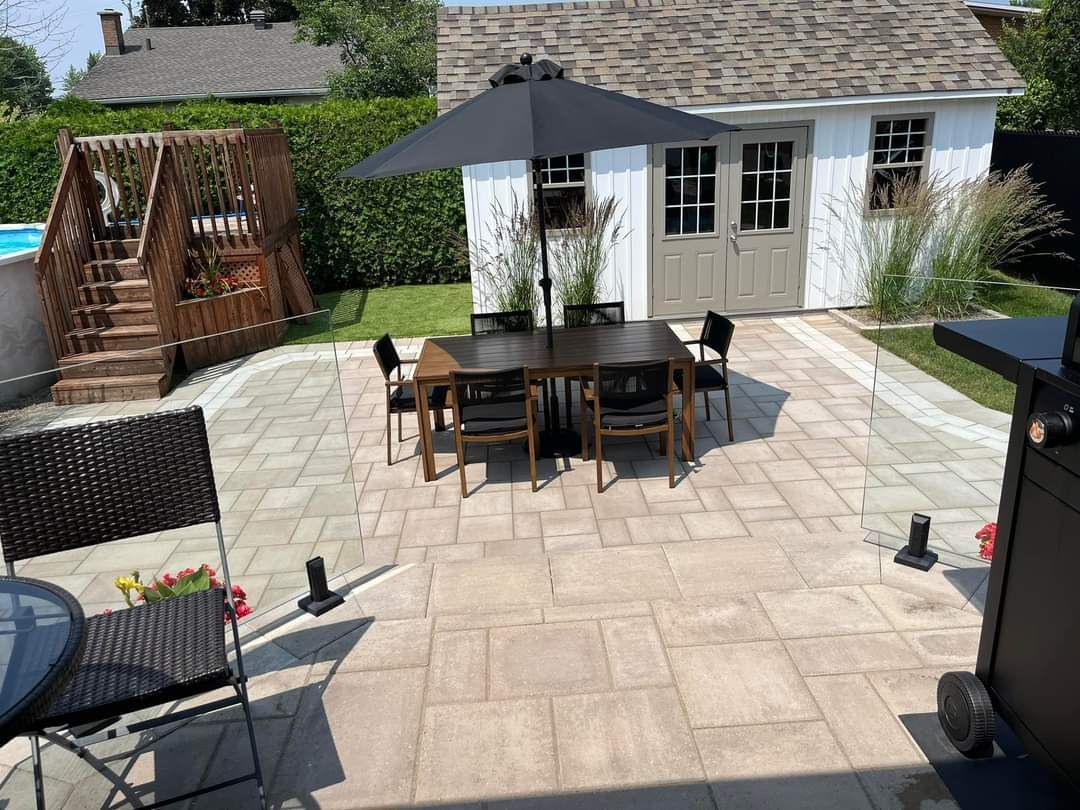 Terrasse avec table, chaises, parasol et abri de jardin. Dalles beiges, pelouse verte et escalier en bois.
