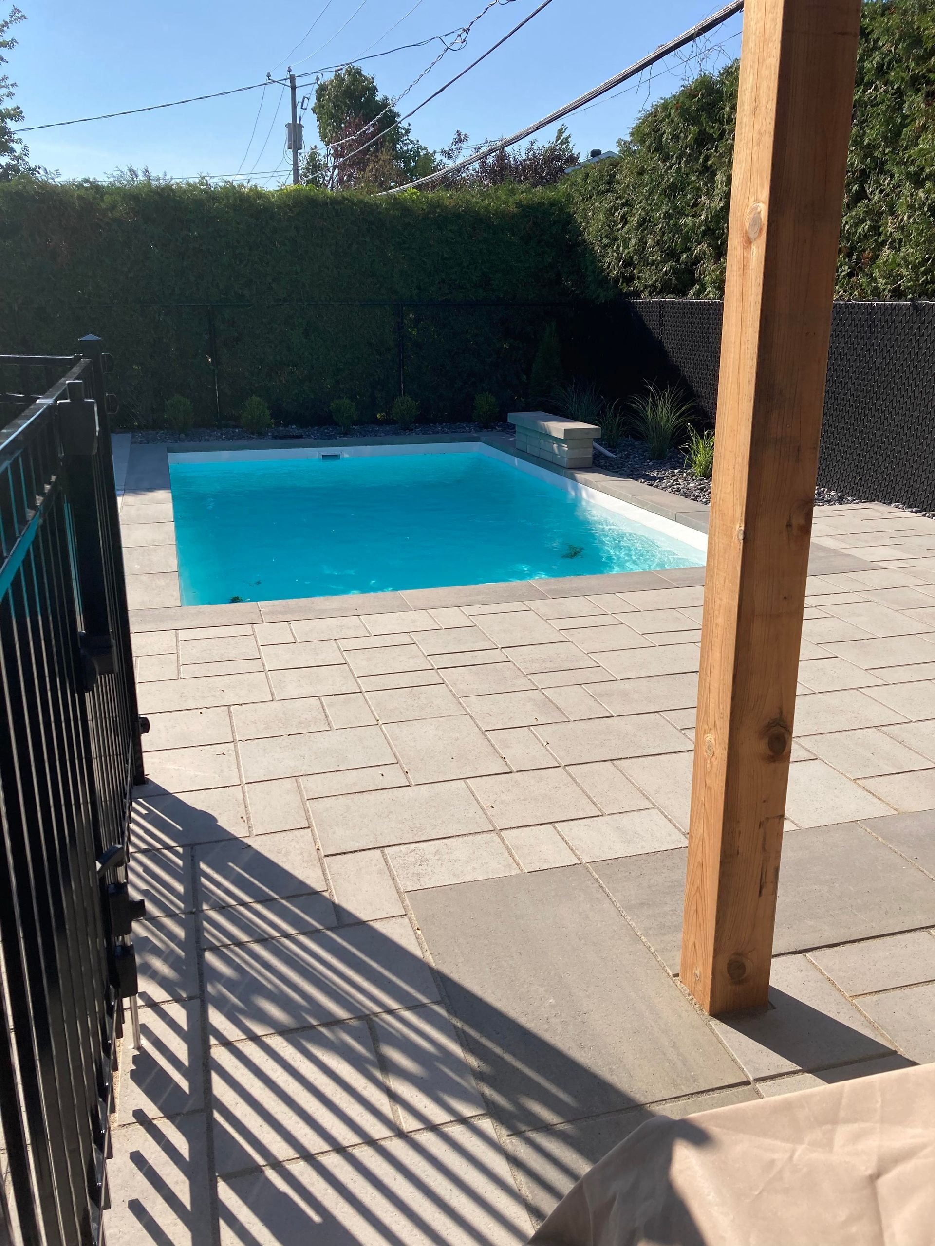 Piscine aux eaux bleu clair entourée de dalles de pierre, d'une poutre de soutien en bois et d'une clôture noire. Haie verte en arrière-plan.