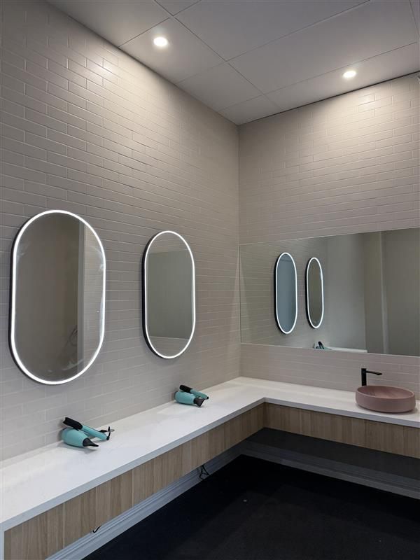 Modern vanity area with two oval backlit mirrors on a tiled wall, a long white counter, and a pink sink with a faucet.
