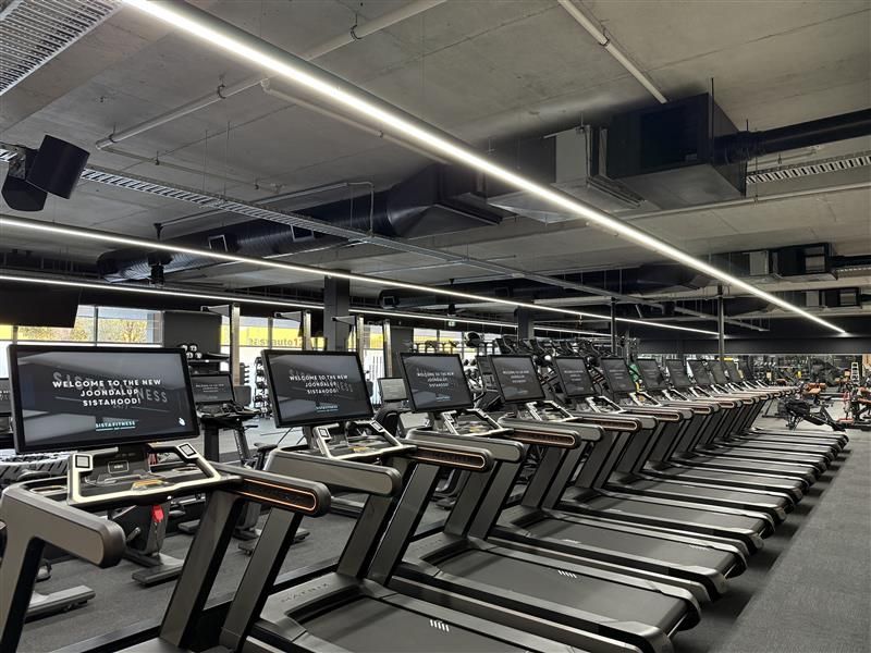 A row of empty treadmills in a modern gym with industrial ceiling lighting and black metal structures.