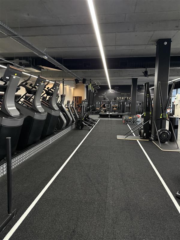 An indoor gym facility featuring a row of cardio machines, a central aisle with floor markings, and overhead lighting.