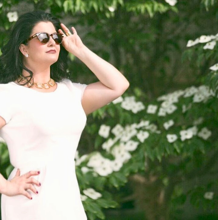 Woman in white dress posing outdoors beside green foliage and white flowers, wearing sunglasses.