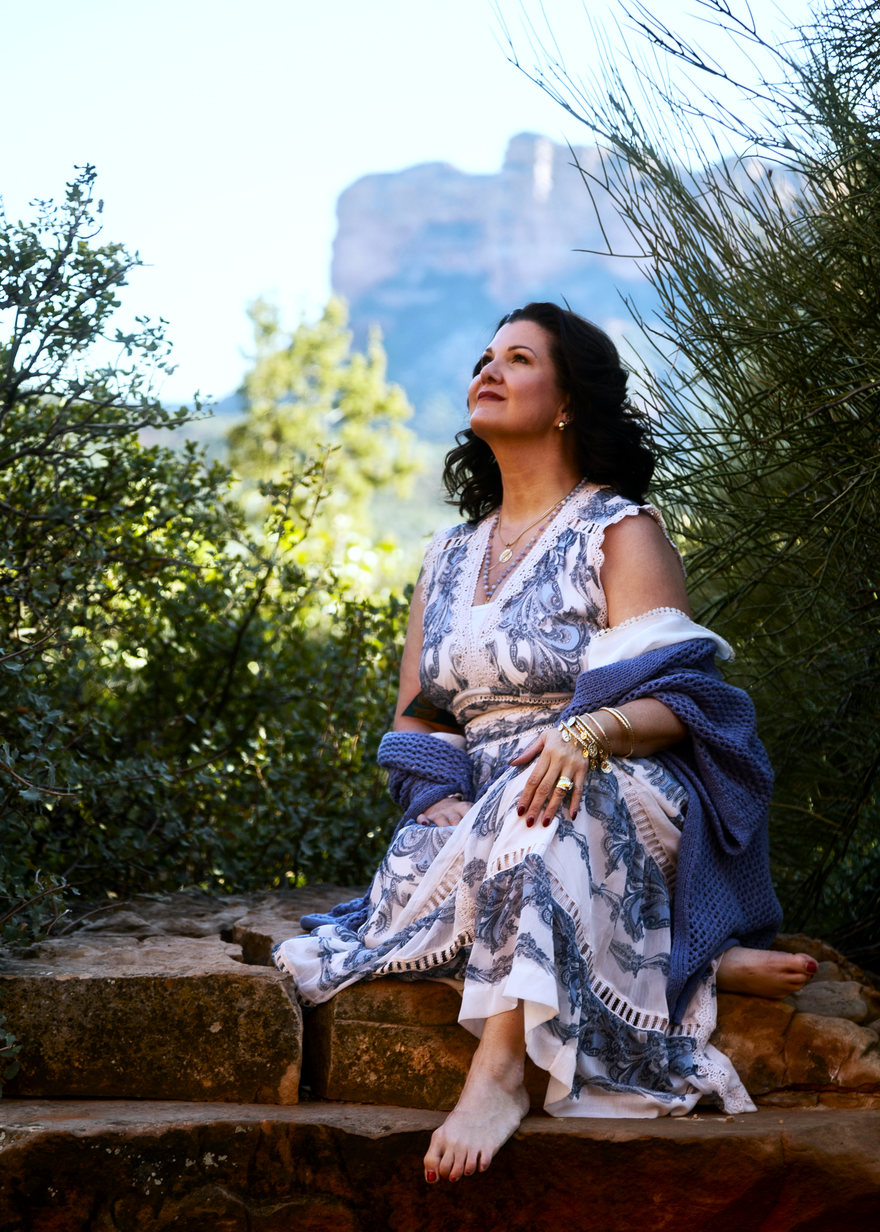 Woman in a floral dress sits on stone steps outdoors, looking upward with trees and a cliff in the background