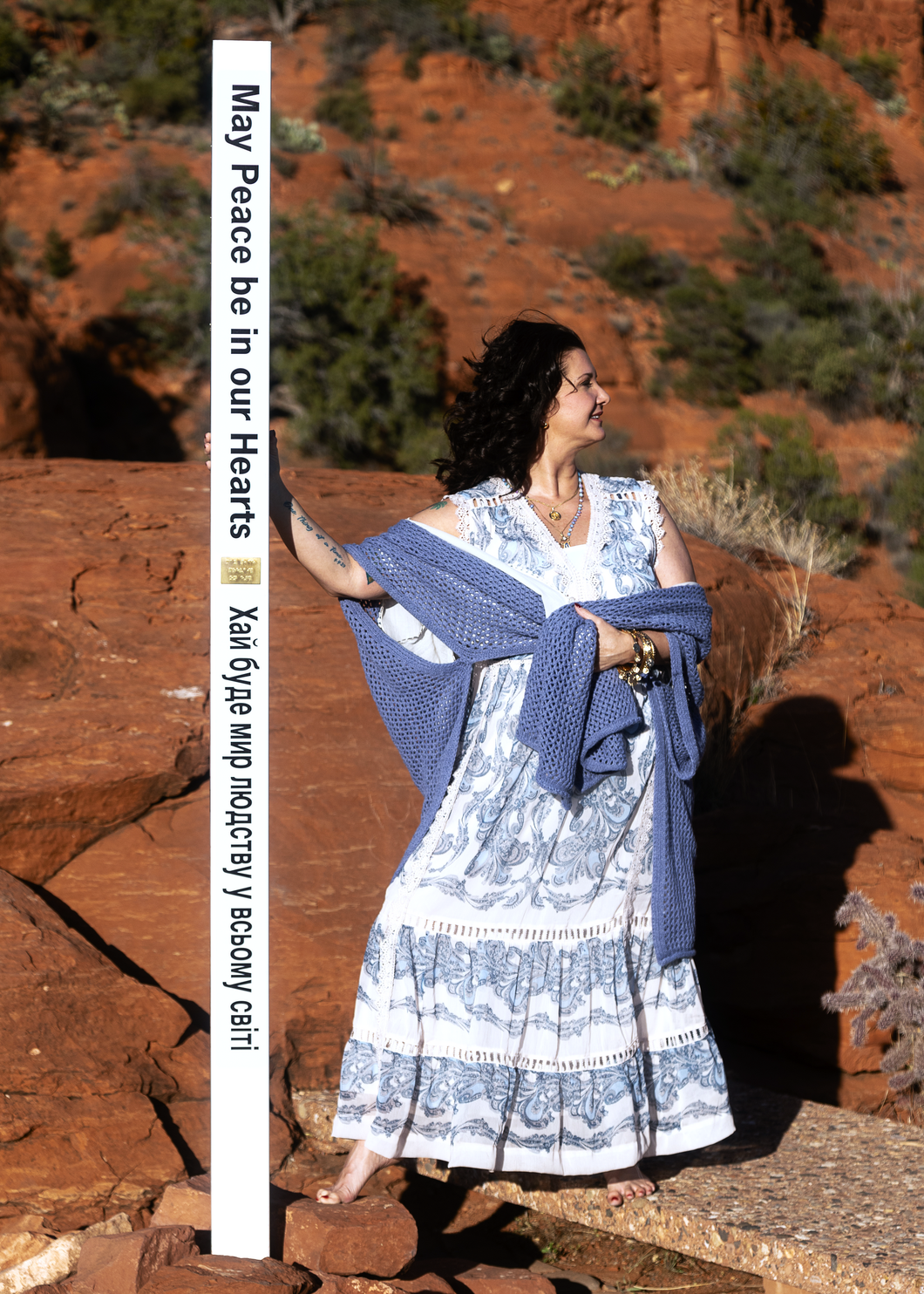 Woman in a blue dress standing on a wooden fence before sunlit red rock cliffs.