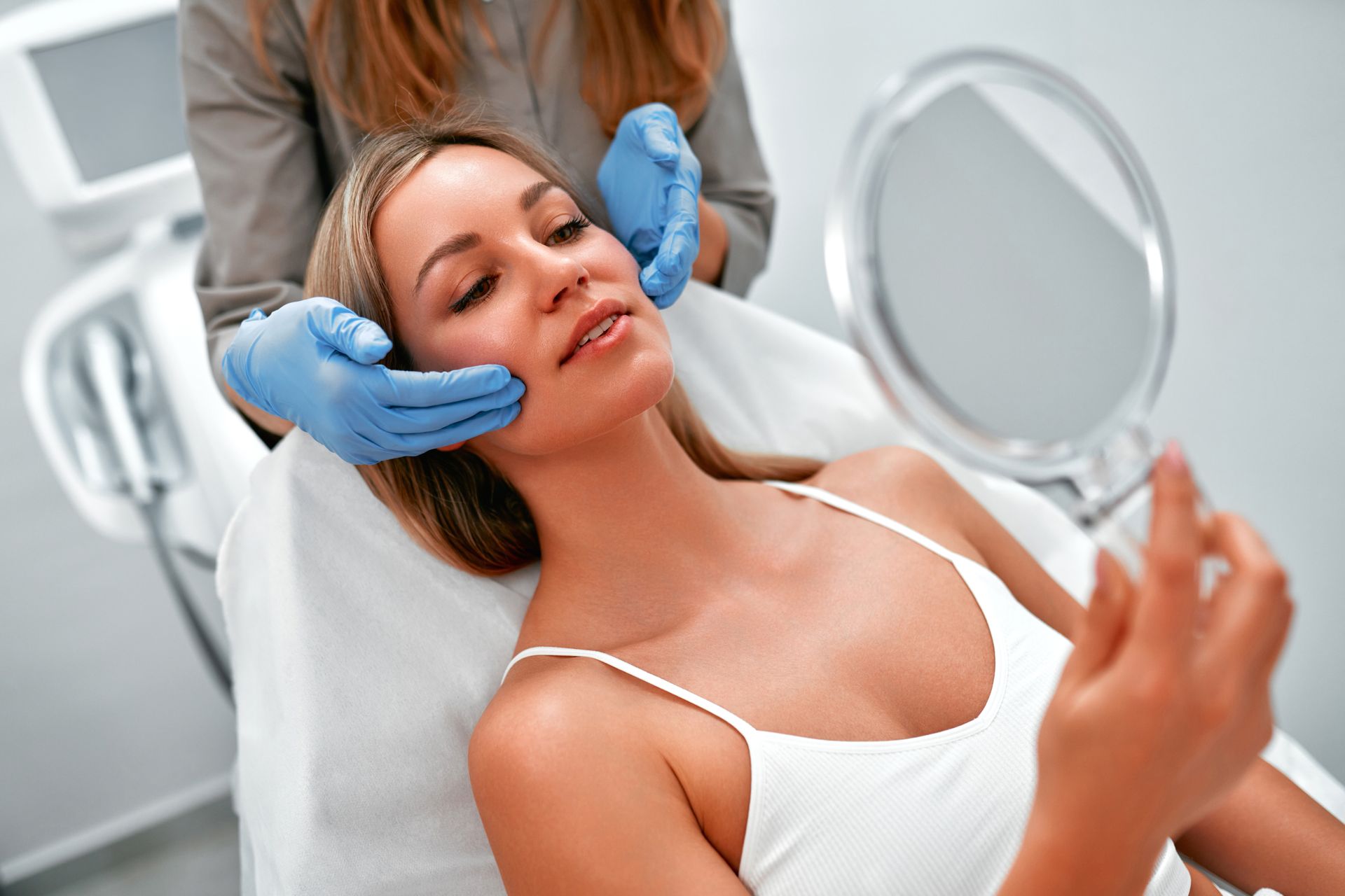 Woman receiving a facial treatment while reclining in a spa chair and looking into a hand mirror