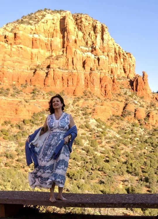 Woman in blue dress standing on a wooden rail before a sunlit red-rock cliff.