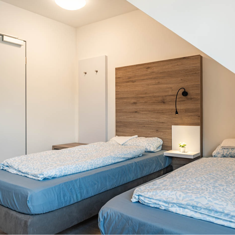 Two beds in a room, with blue linens, wooden headboard, and bedside tables.