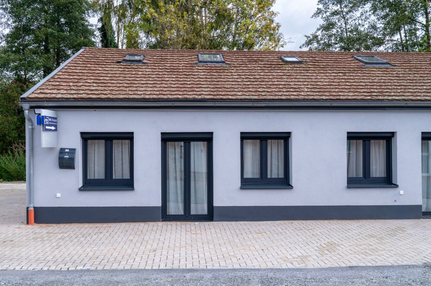 Gray building with black windows and door, brown roof, and brick paving.