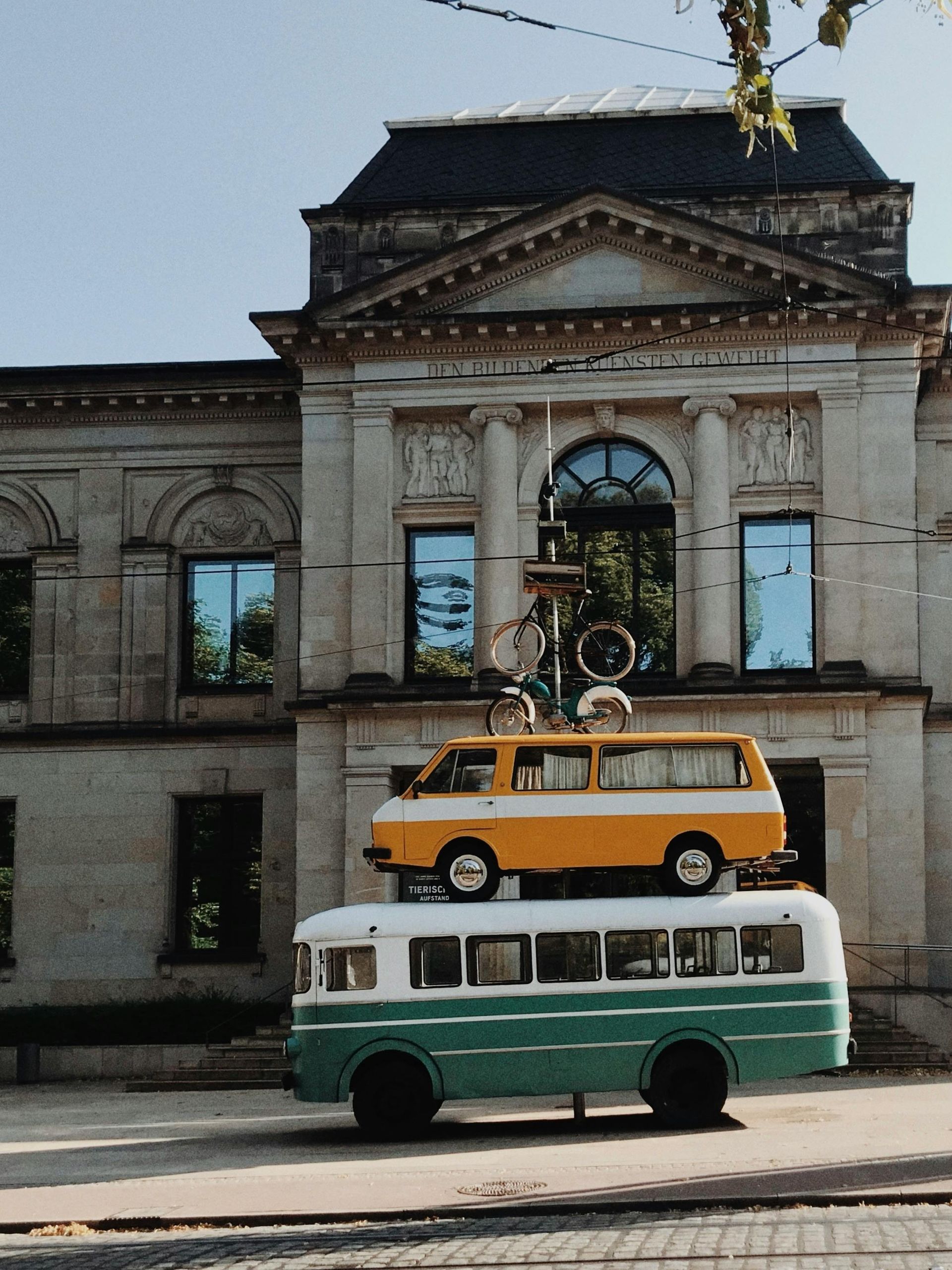 Green and white bus with yellow van stacked in front of a classical building with blue windows.