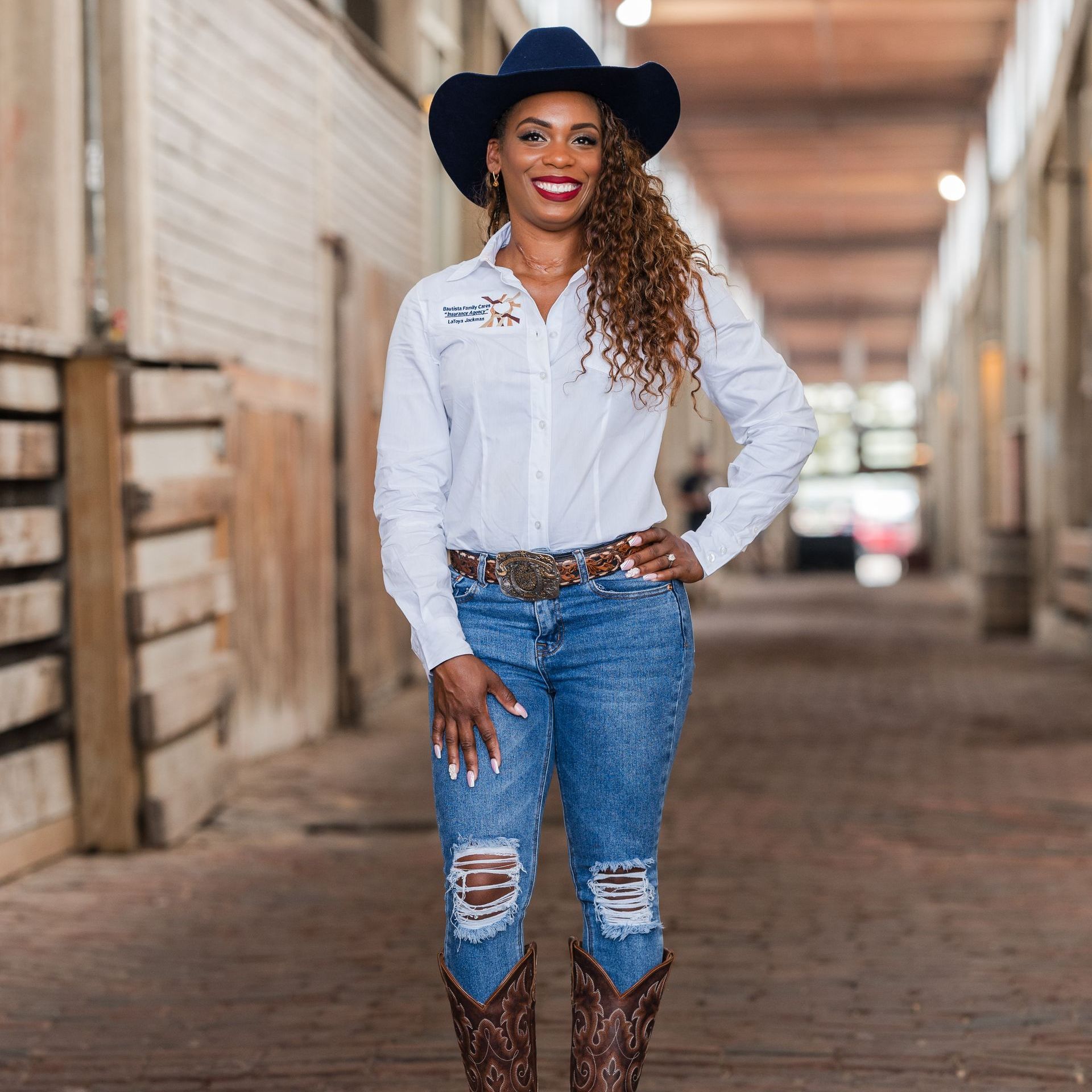 Woman in white shirt, blue jeans, and cowboy hat standing in a barn aisle.