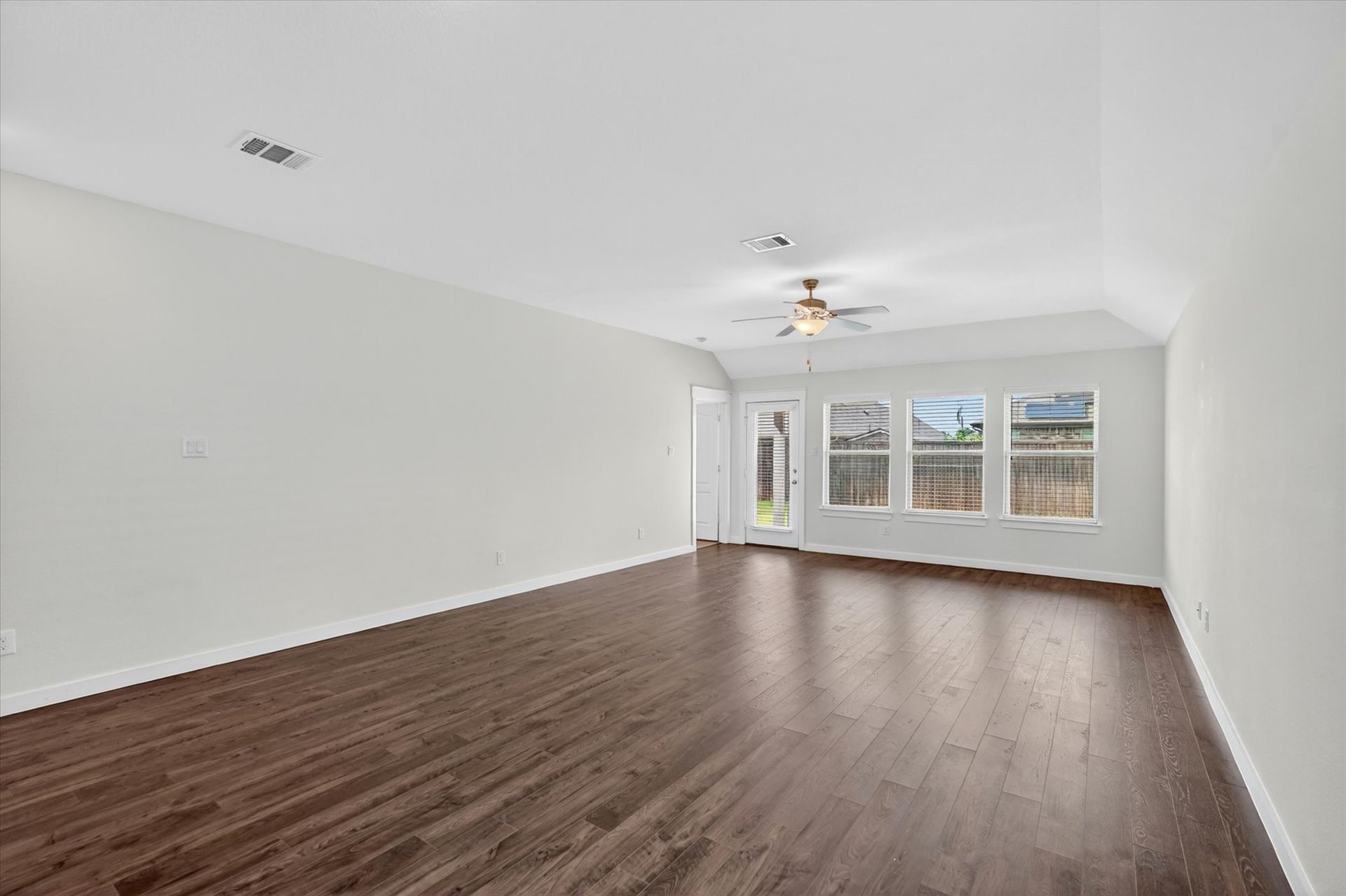 Empty living room with dark wood floors, white walls, and large windows with blinds.