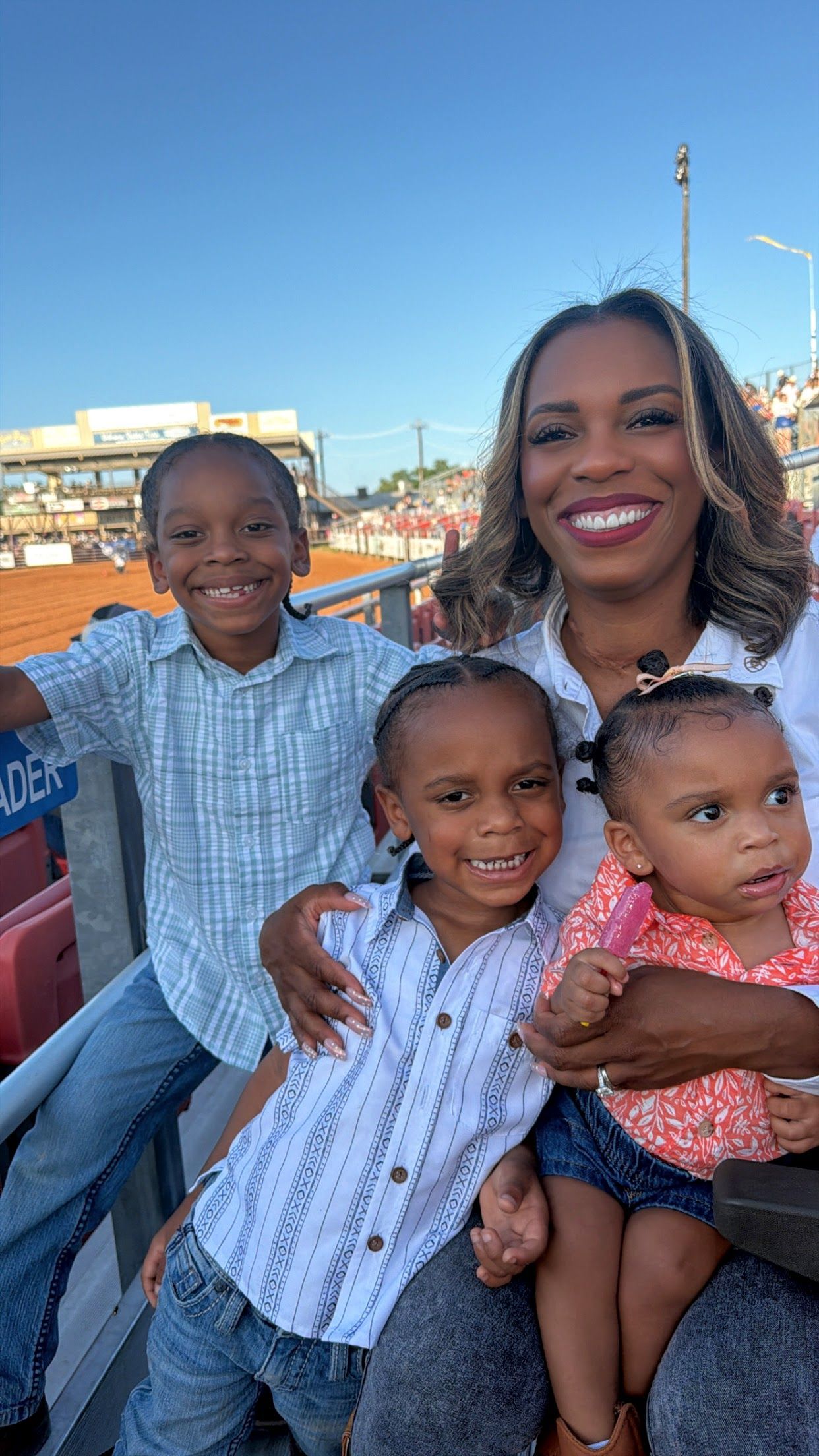 Woman and three children smiling outdoors in front of a waterfront and blue sky.