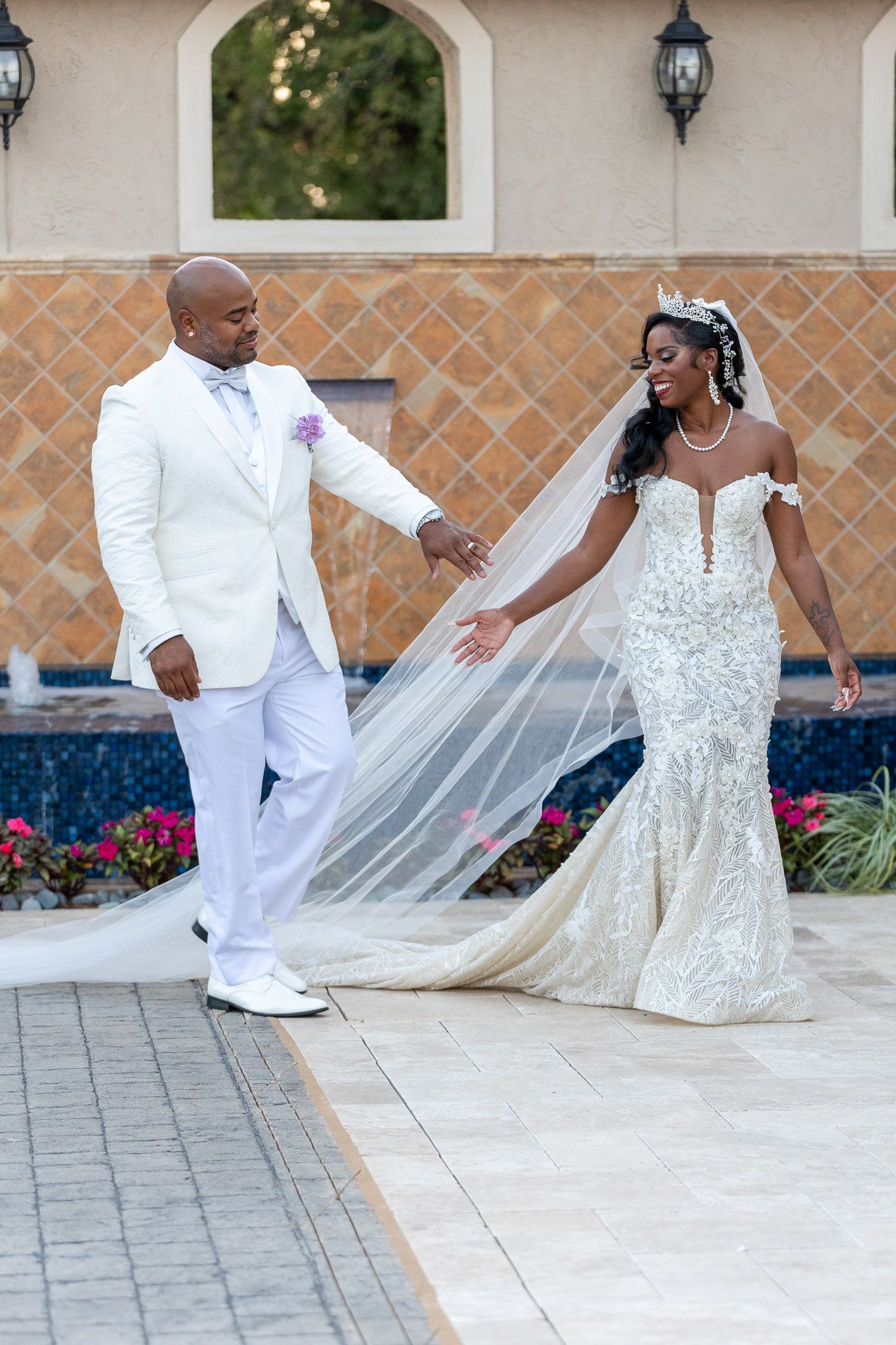 Couple in white wedding attire holding hands outdoors, bride’s veil trailing on a tiled walkway.