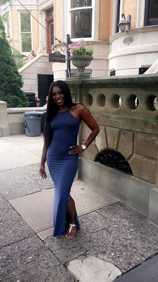 Woman in a blue dress posing on a city sidewalk beside a stone building.