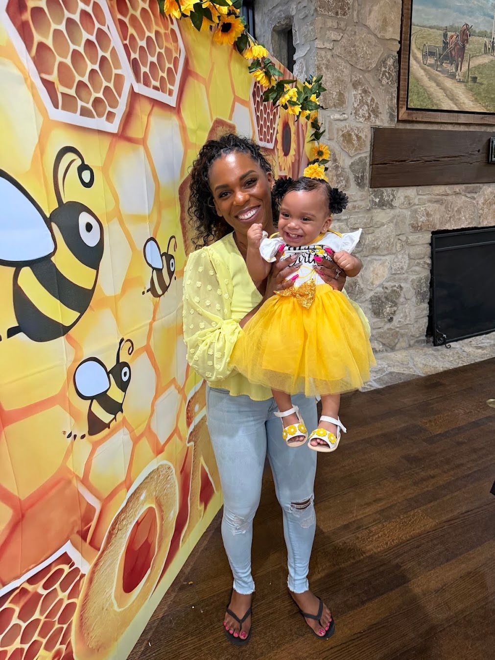 Woman holding a baby in yellow dresses beside a honeycomb mural in a room with dark wood floors